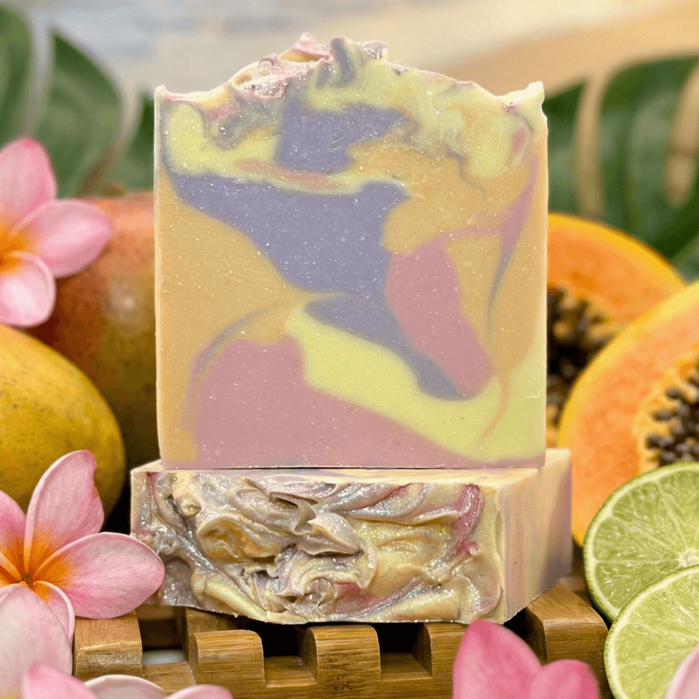 Handcrafted soap bar with marbled swirls of yellow, lavender, and pink throughout. Displayed on bamboo soap dish surrounded by plumeria flowers, fresh papaya, mango, and lime slices.