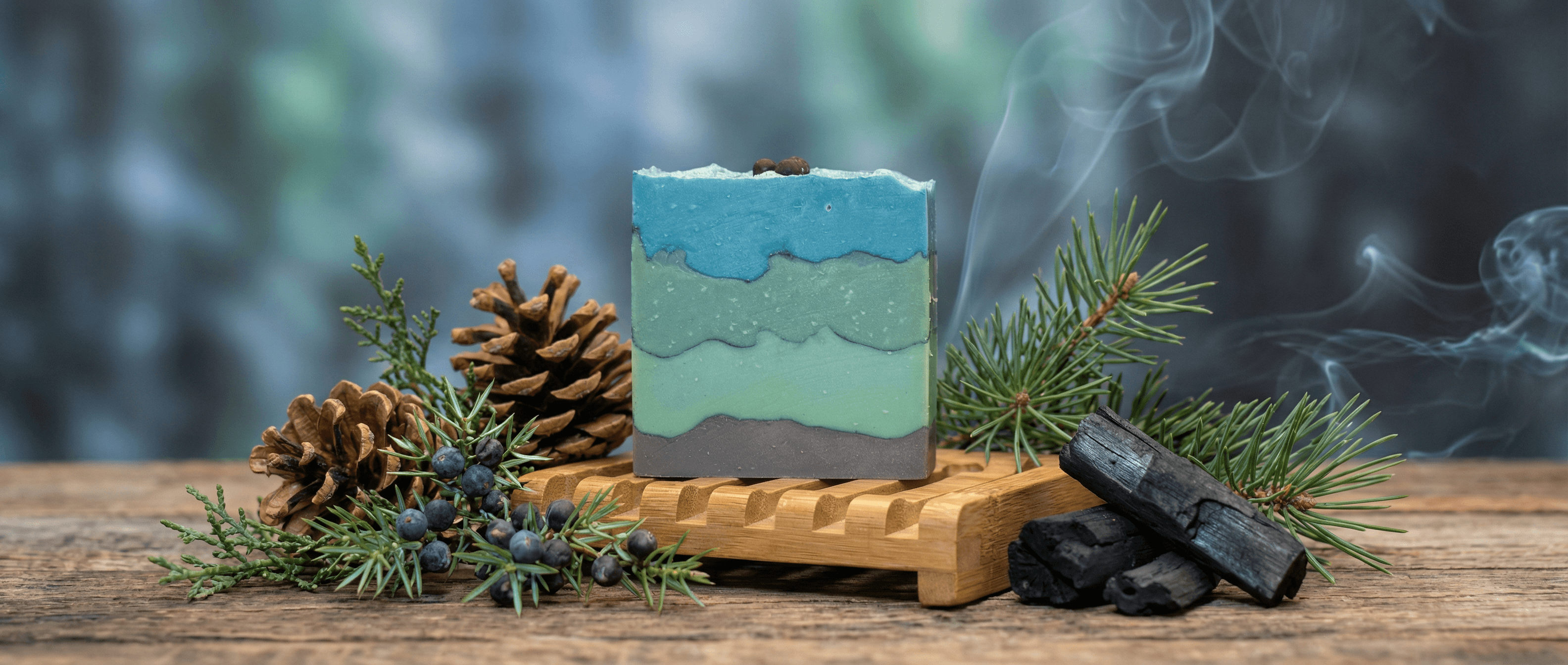 Layered handmade soap bar in ocean blue, seafoam green, and charcoal gray stripes, displayed on wooden soap dish with pine branches, pinecones, juniper berries, and charcoal pieces.