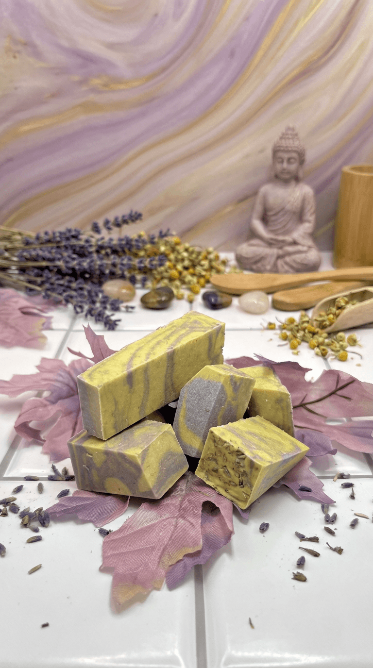 Yellow and gray marbled soap bars with swirled pattern, displayed on purple silk fabric. Surrounded by dried lavender stems, chamomile flowers, polished stones, and a Buddha statue.