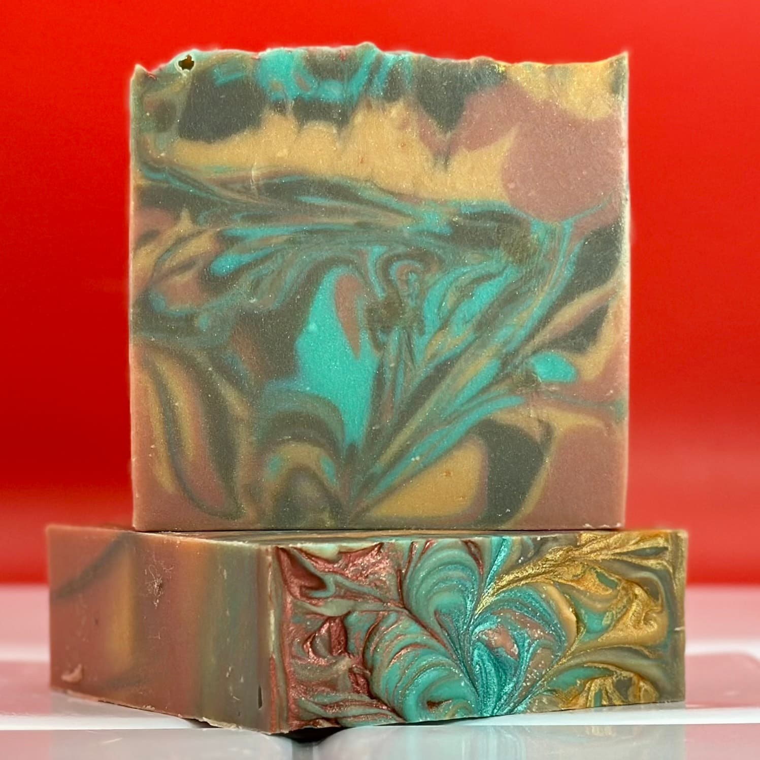 Two stacked handcrafted soap bars with turquoise, gold, and mauve swirled patterns creating feather-like designs on an orange-red background. Cold process soap with marbled surface texture.