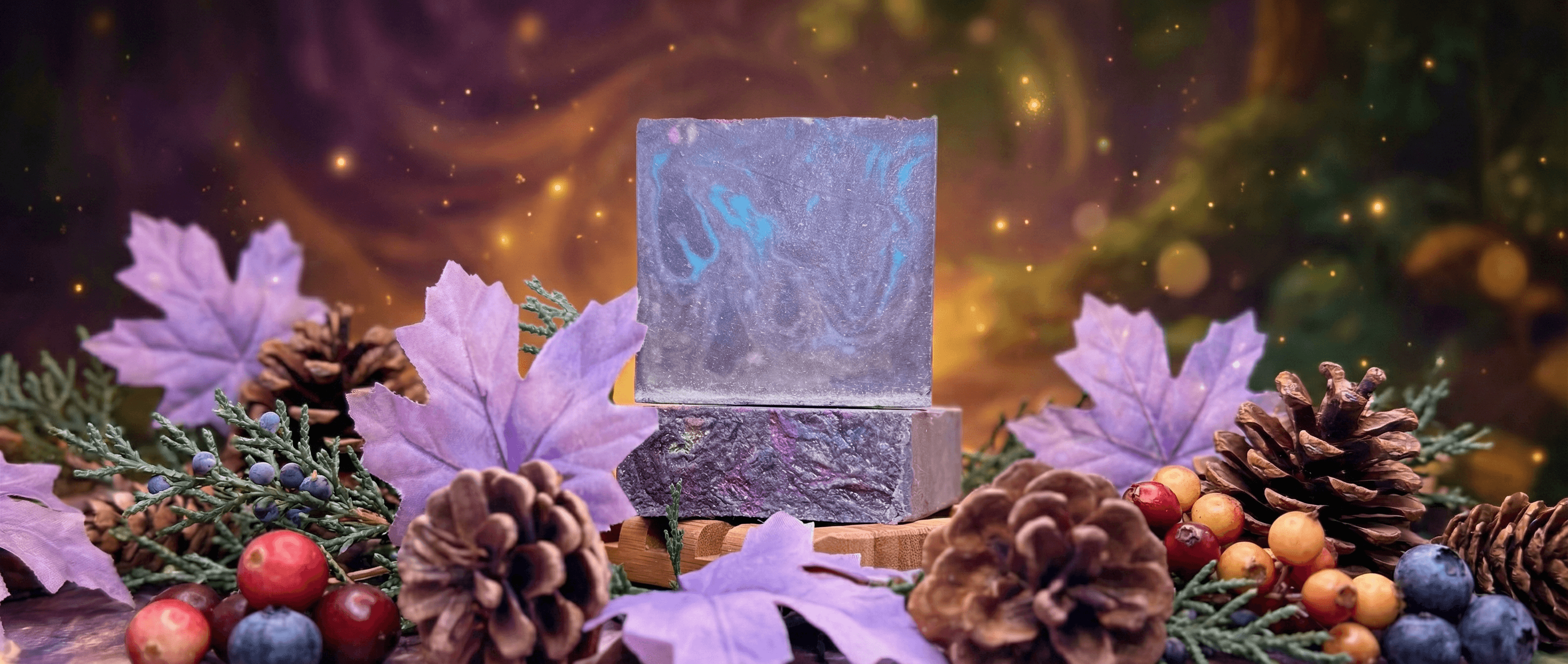 Two stacked bars of purple and blue swirled soap with marbled texture, displayed on wooden soap dish surrounded by pine cones, purple leaves, evergreen sprigs, and colorful berries against a dreamy bokeh background.
