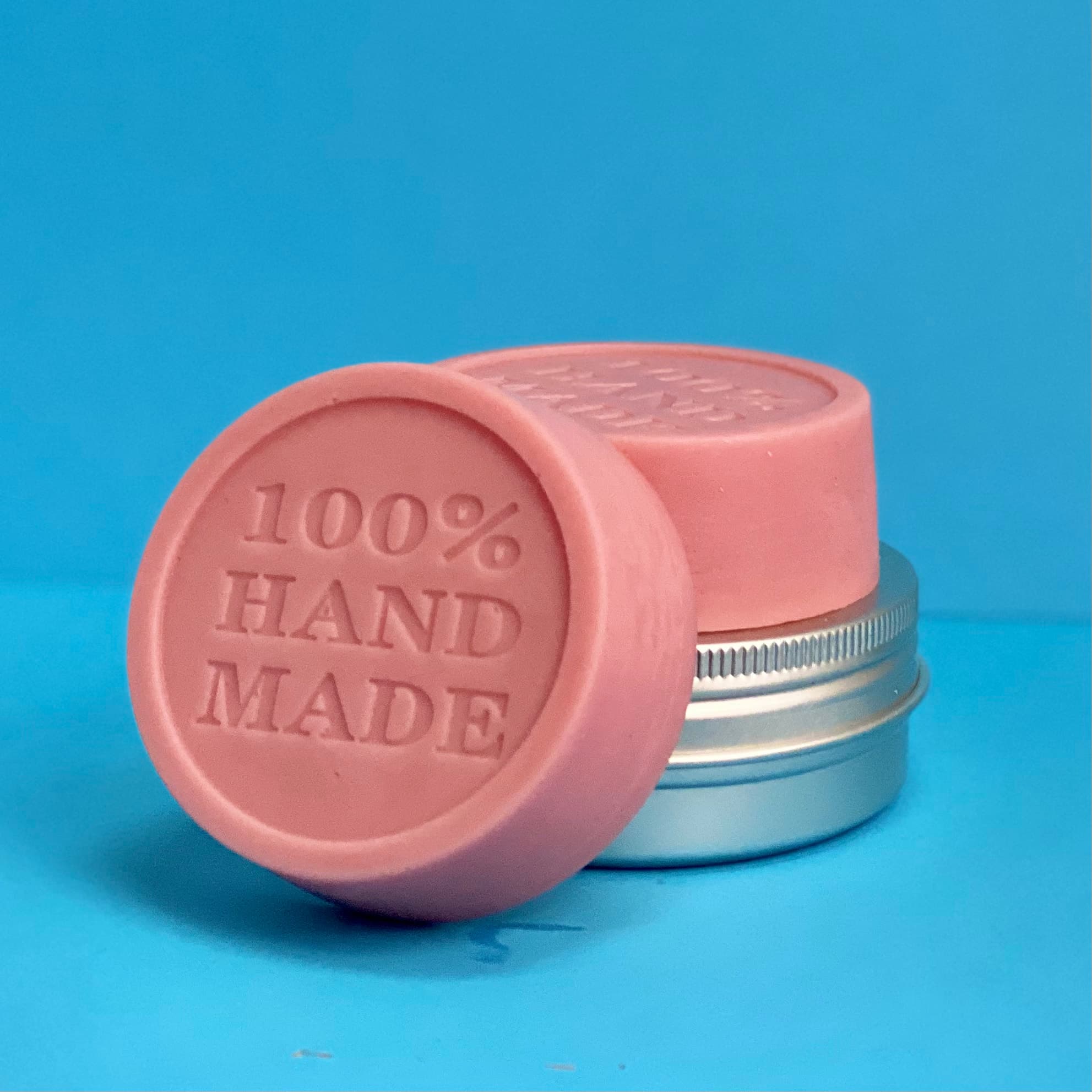 Two round pink solid lotion bars with embossed '100% HAND MADE' text on top, displayed against a bright blue background with a silver tin container.