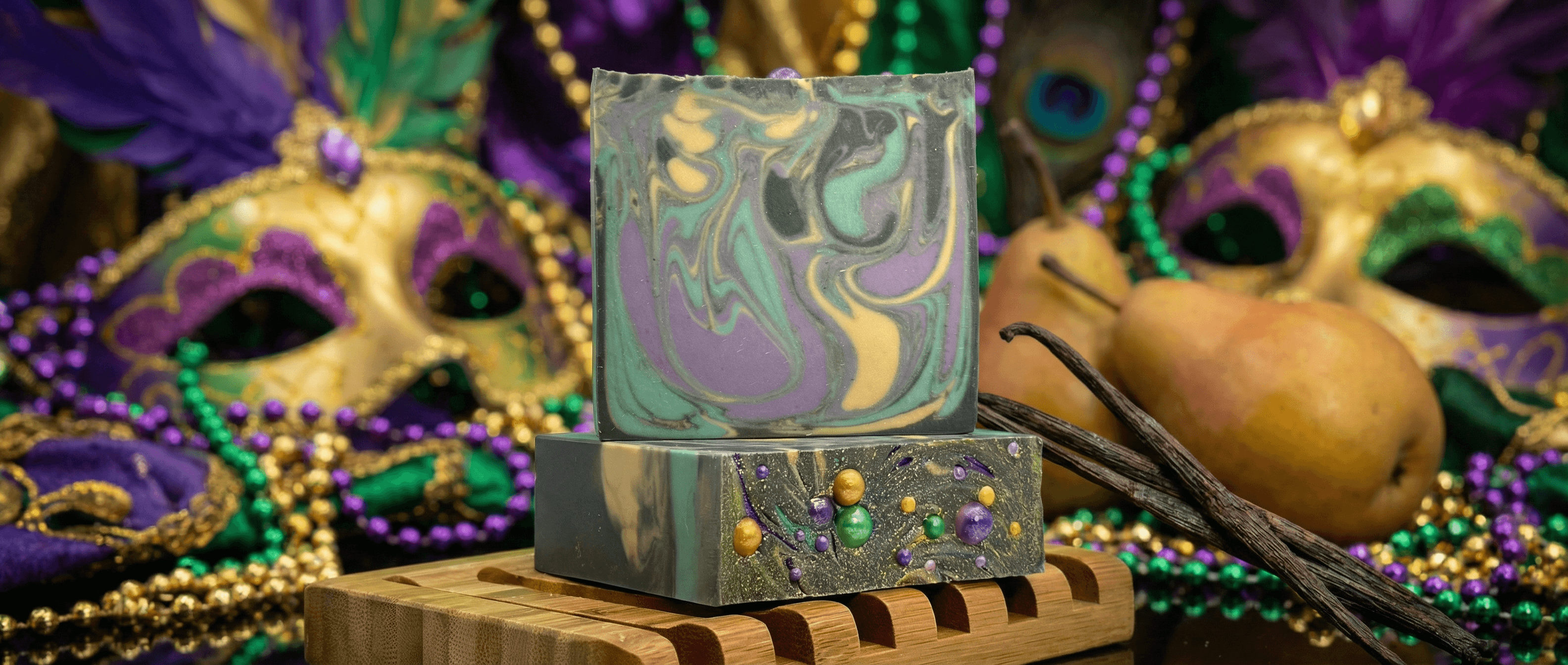 Handcrafted soap bar with swirled purple, green, gold, and gray marbling on top. Bottom half features green base embedded with purple, gold, and green spherical beads, displayed on wooden soap dish with vanilla beans and Mardi Gras decorations.