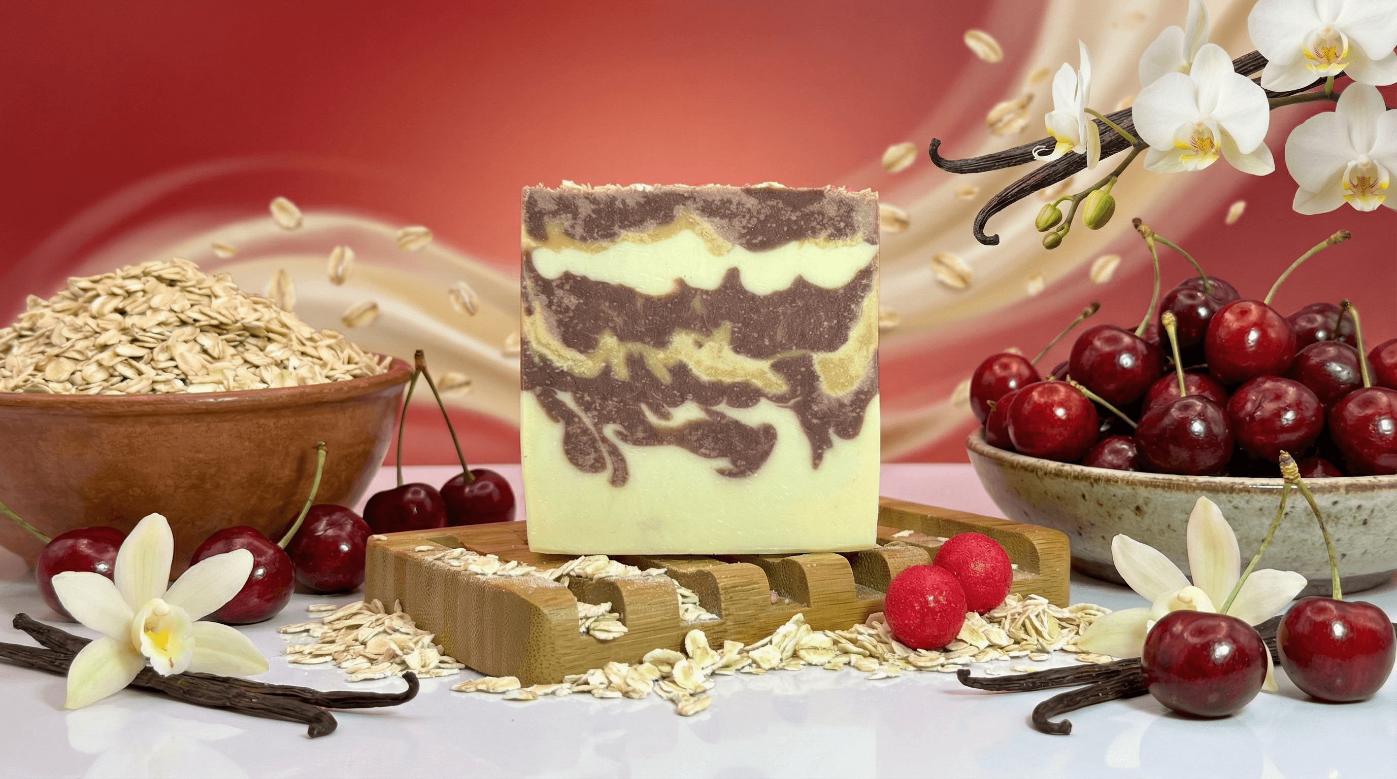 Handcrafted bar with swirled brown and pale yellow layers creating a marbled pattern. Displayed on bamboo tray surrounded by oats, fresh cherries, vanilla beans, and white orchid flowers.