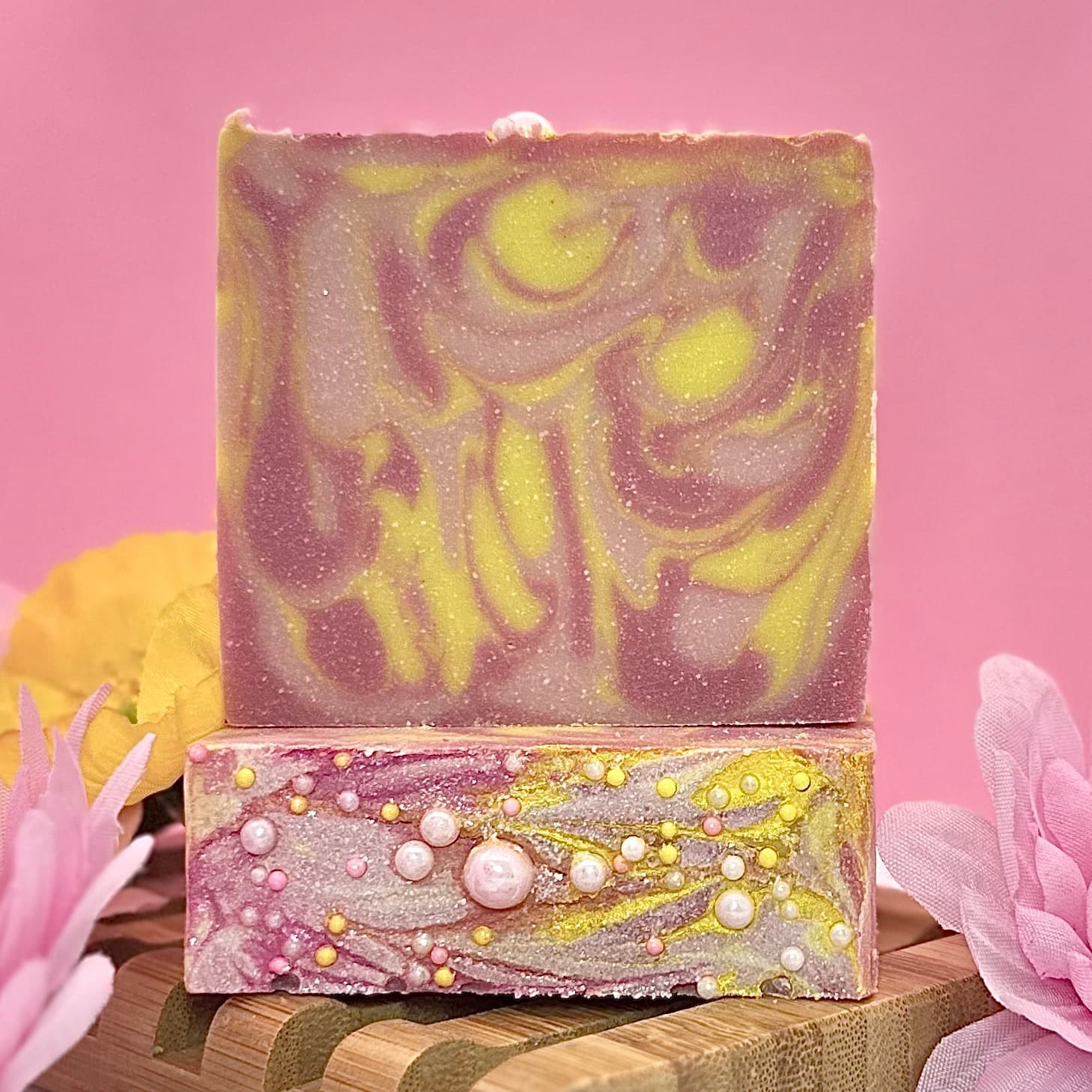 Two stacked handmade soap bars with purple and yellow marbled swirls. Bottom bar features pearl embellishments, top bar has swirled pattern. Displayed on wooden stand with pink flowers.