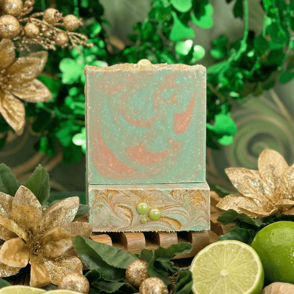 Rectangular handcrafted soap bar with mint green and coral swirled top, resting on wooden soap dish. Surrounded by gold poinsettias, green shamrocks, and fresh limes.
