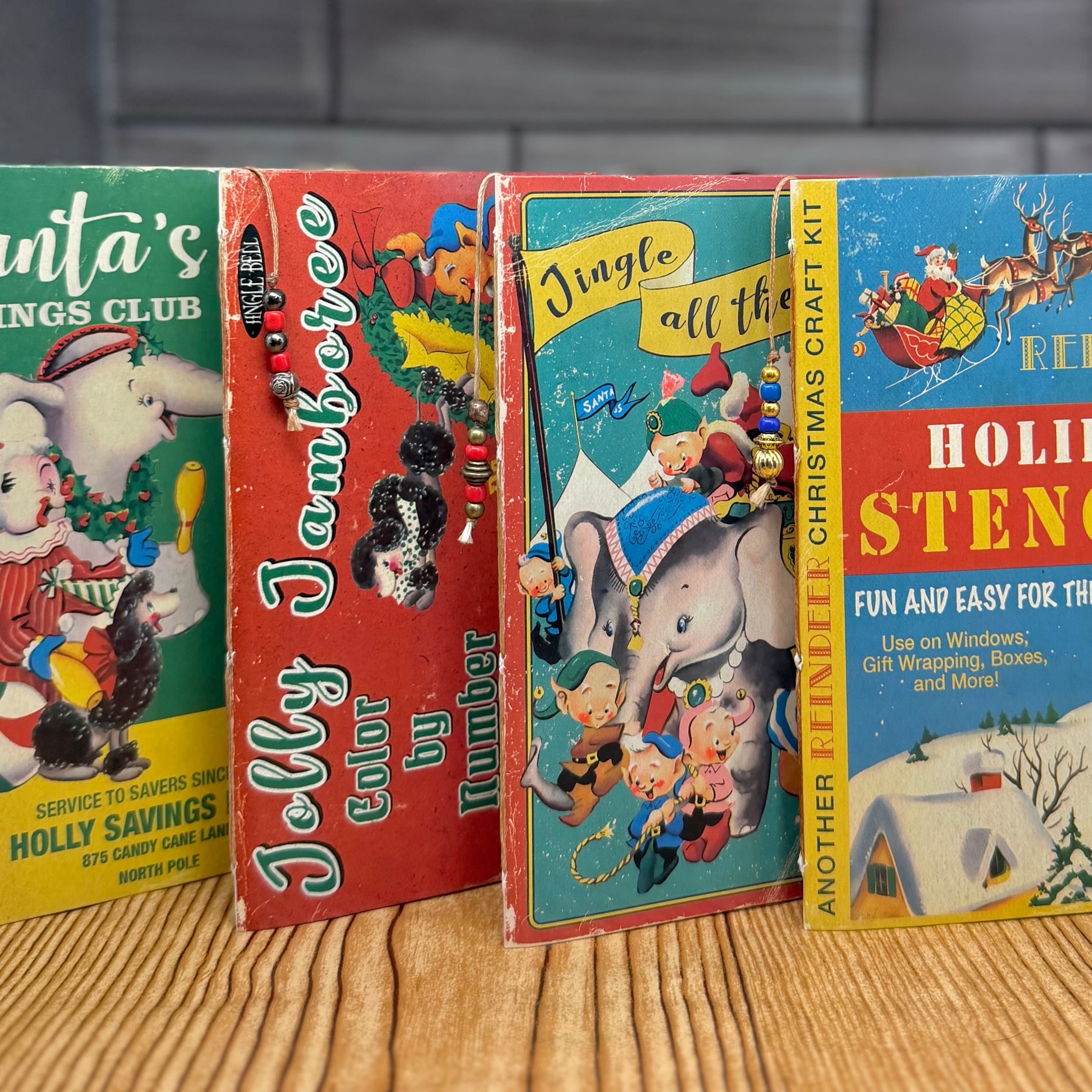 Four vintage-style Christmas card journals standing upright on wood surface, featuring retro holiday illustrations including Santa, reindeer, elephants, and poodles in red, green, blue and yellow.