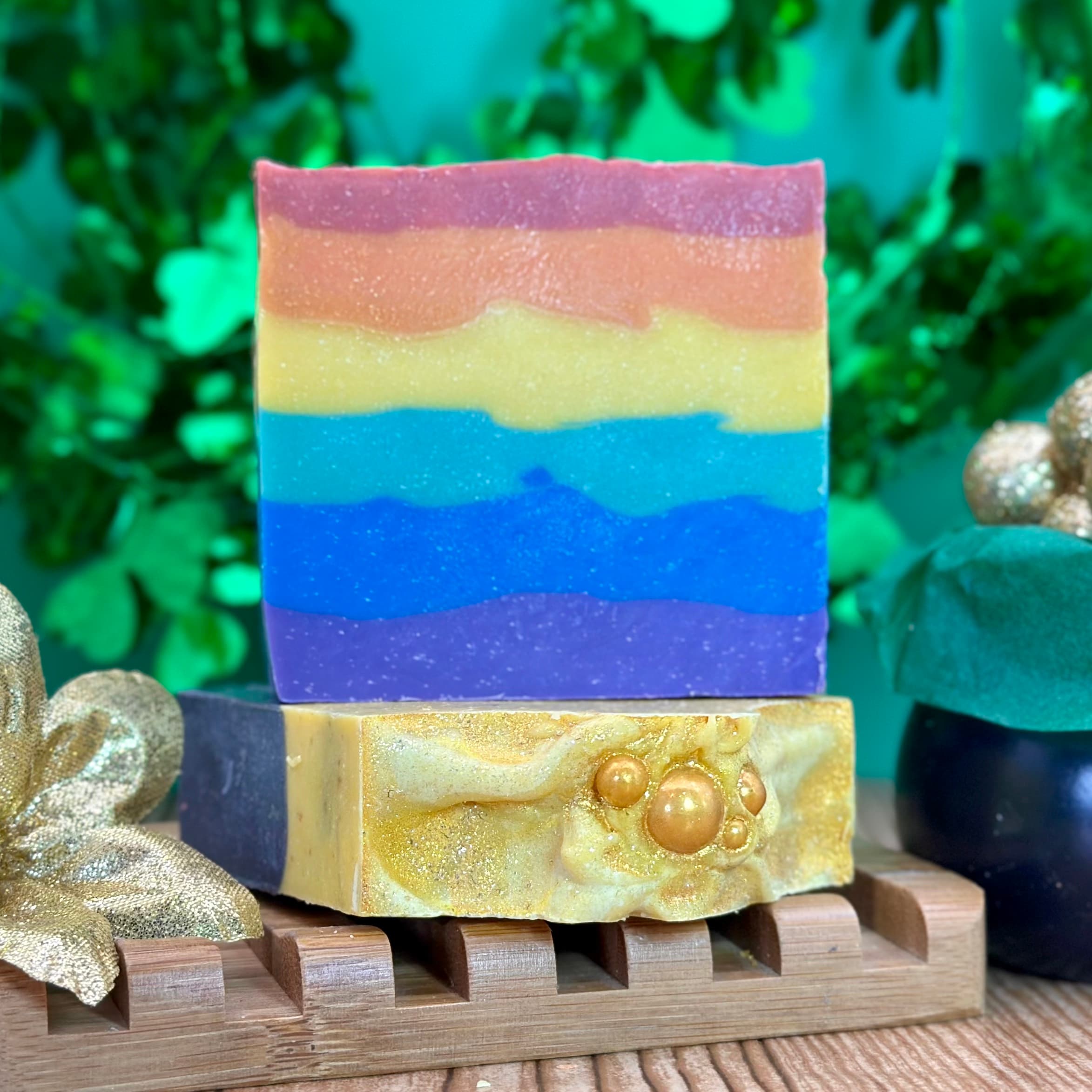 Two stacked handcrafted soap bars on a wooden soap dish with gold ribbon. Top bar features rainbow stripes from pink to purple with shimmer. Bottom bar is golden yellow with swirled texture and pearl embellishments.