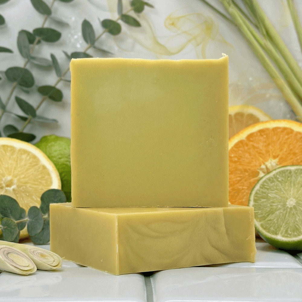 Pale yellow rectangular soap bars stacked on white surface, surrounded by fresh citrus slices, eucalyptus sprigs, lemongrass stalks, and sliced onion. Smooth, minimal appearance with bright lifestyle styling.