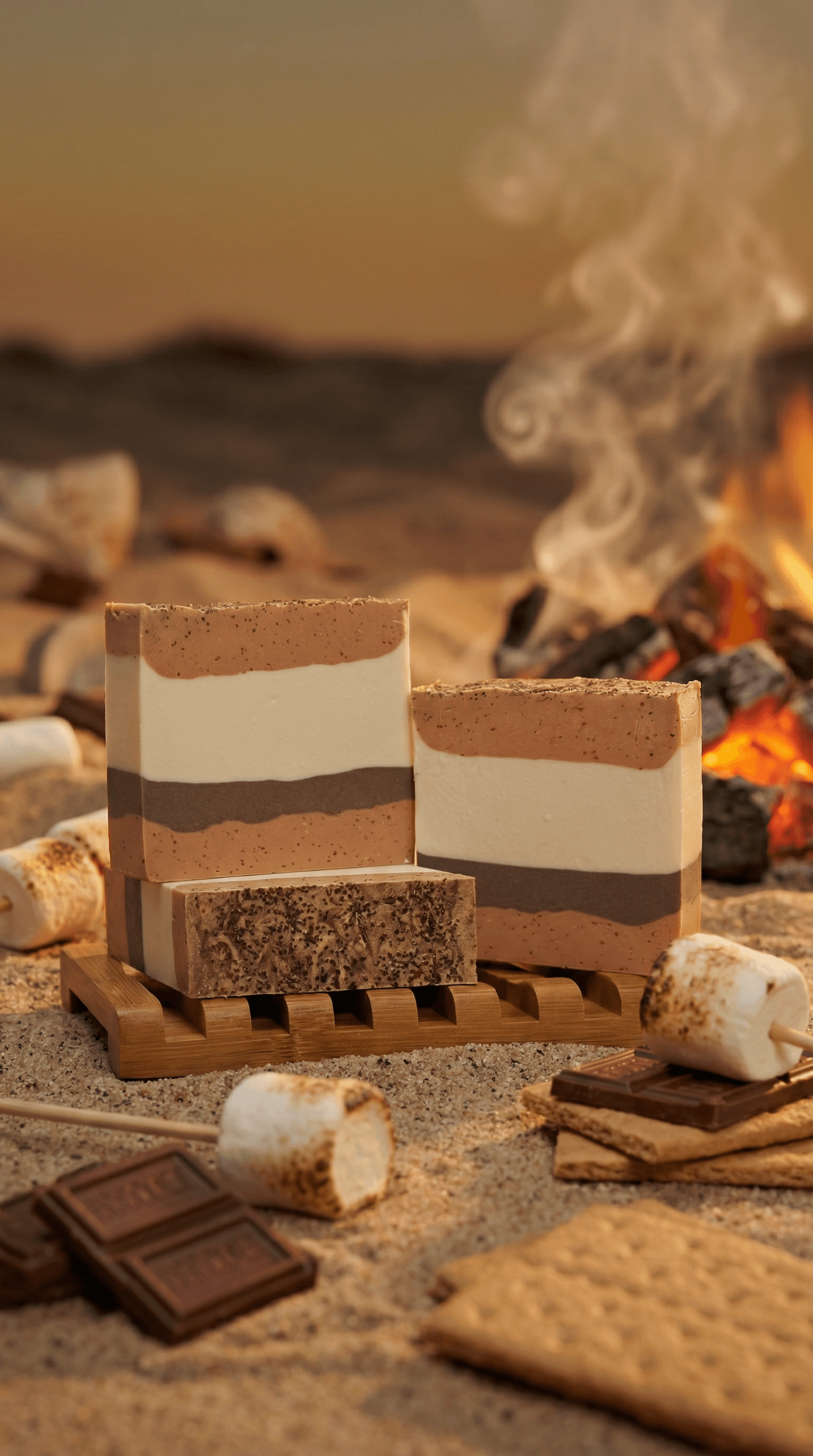 Three layered bars of s'mores-inspired soap with brown, cream, and tan stripes resting on a wooden soap dish, styled with marshmallows, chocolate, and graham crackers beside a campfire.