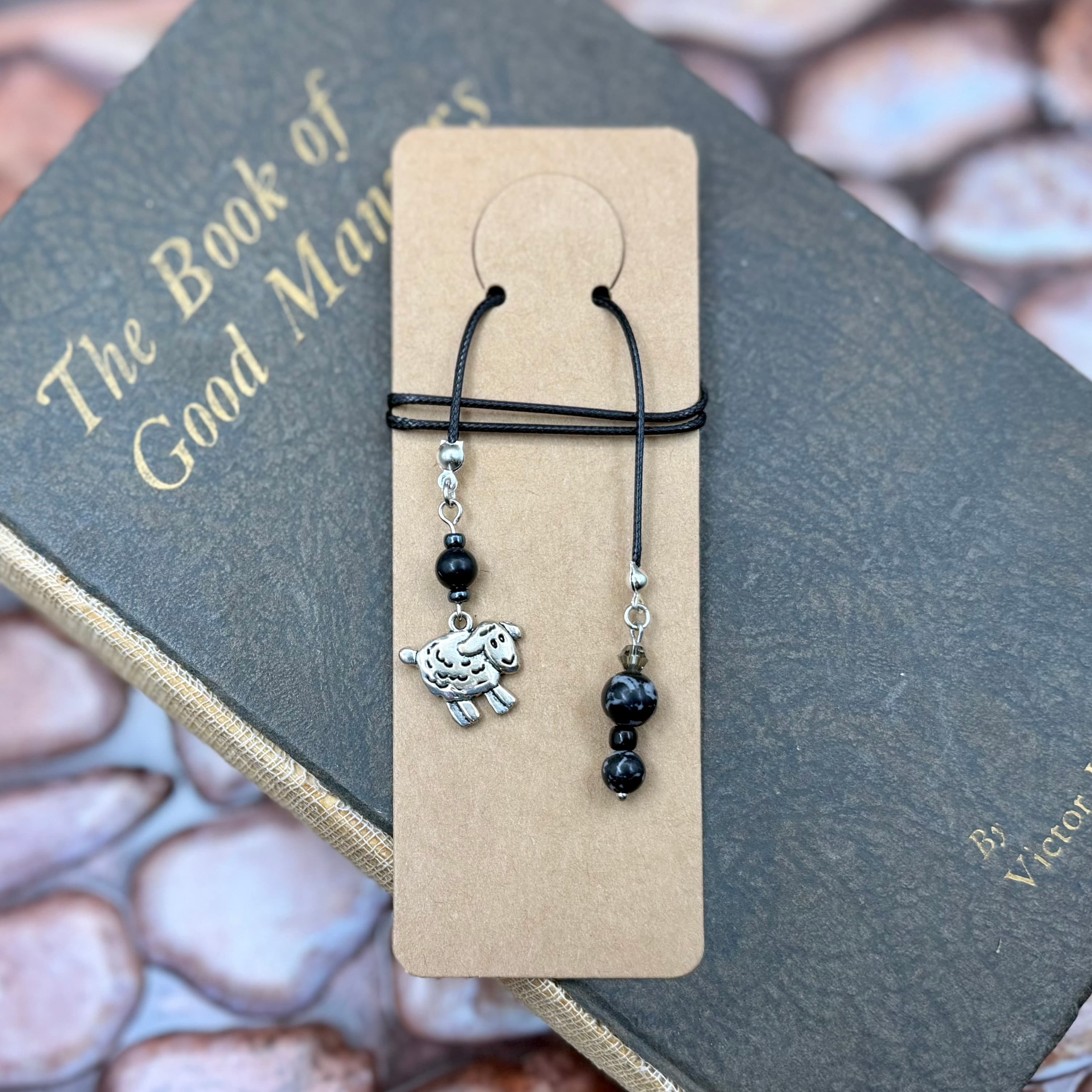 Handmade bookmark with black cord and beads featuring a silver sheep charm, displayed on a tan card atop a gray vintage book titled 'The Book of Good Manners'.