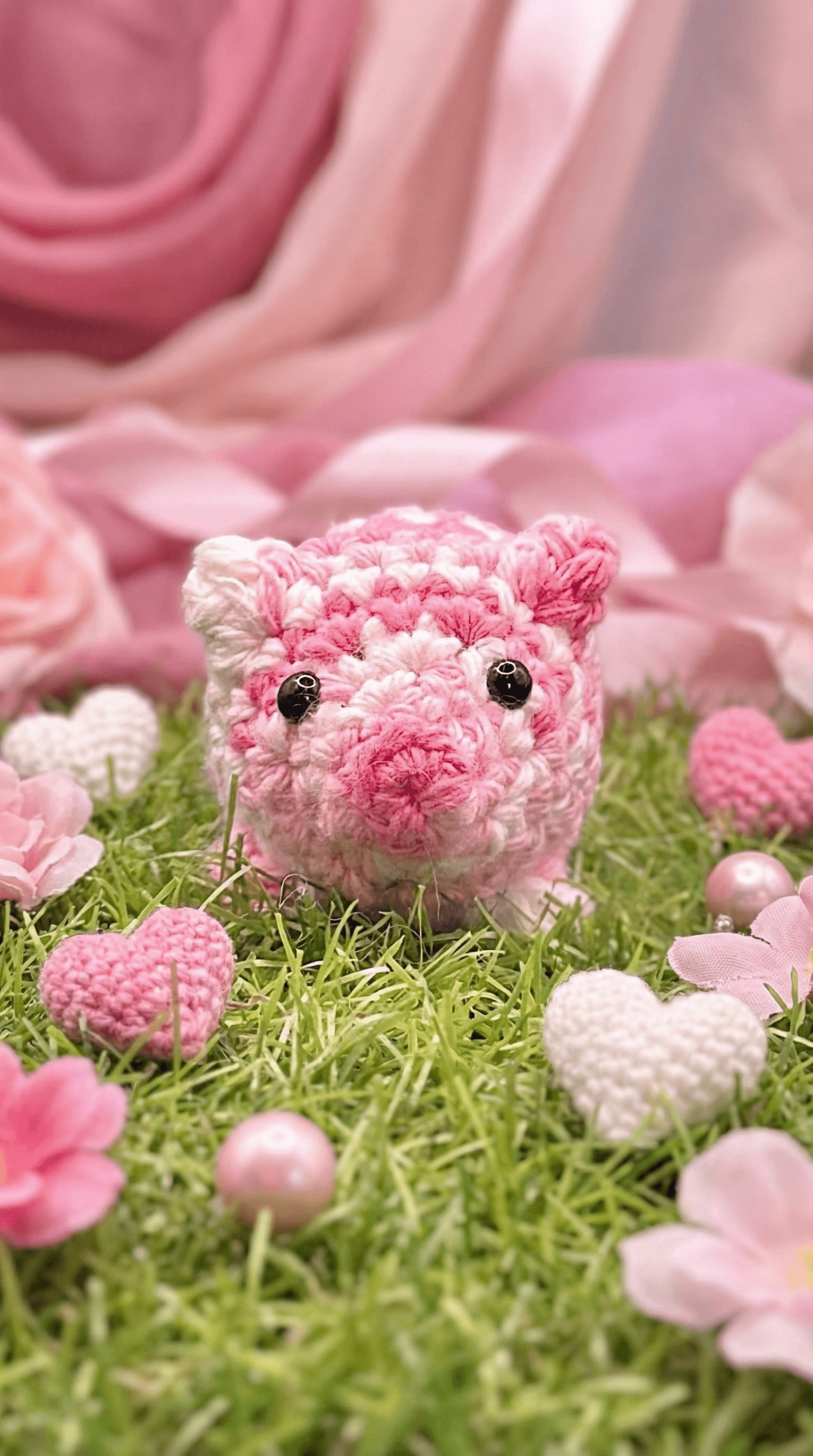 Hand-crocheted pink and white variegated pig-shaped sachet with black bead eyes, displayed on artificial grass surrounded by pink fabric flowers, hearts, and rose petals.