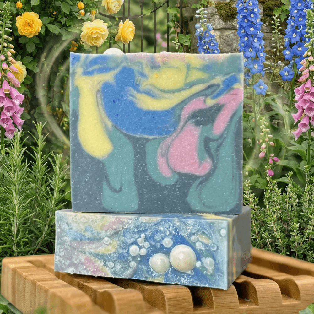 Two handcrafted soap bars on wooden rack against garden backdrop. Front bar shows swirled blues, yellows, and pinks; bottom bar features pearl embeds and speckled texture.