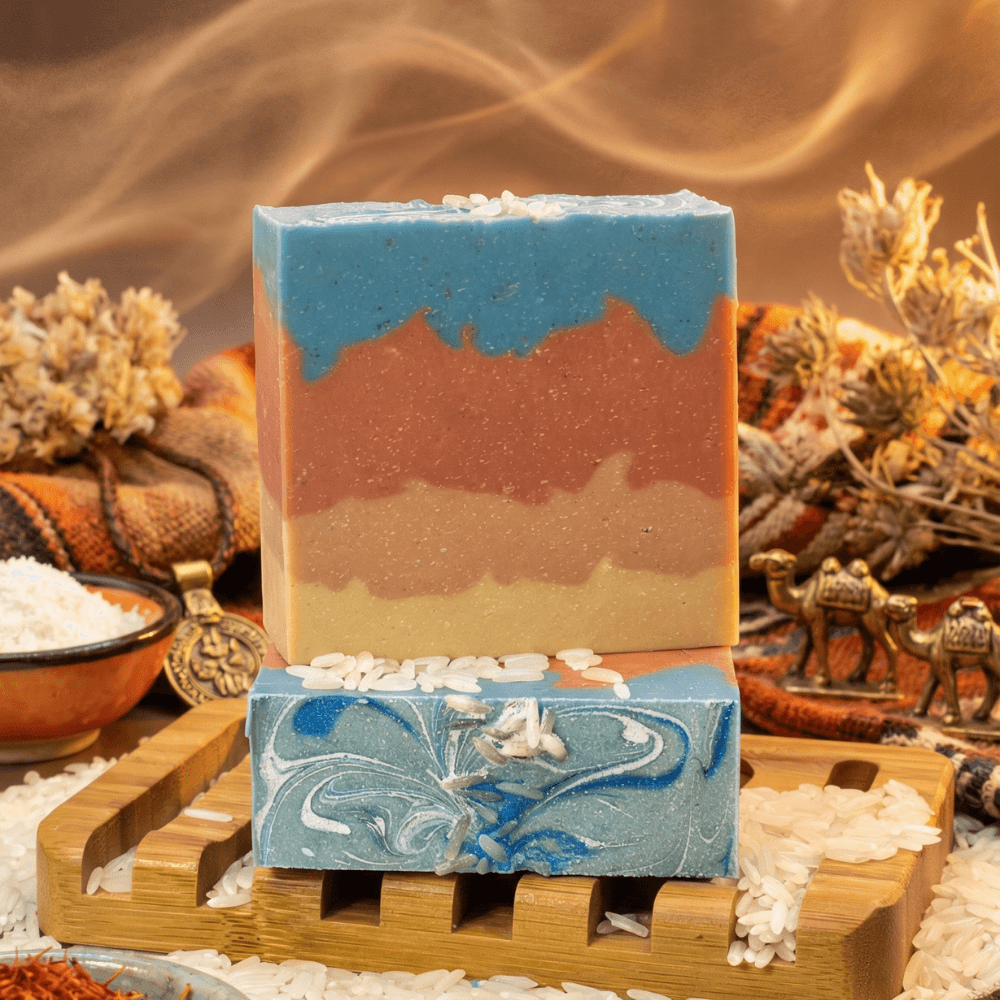 Two stacked handmade soap bars on a wooden tray with white soap flakes. Top bar features layered desert colors in blue, rust, and tan. Bottom bar shows swirled blue and white pattern.