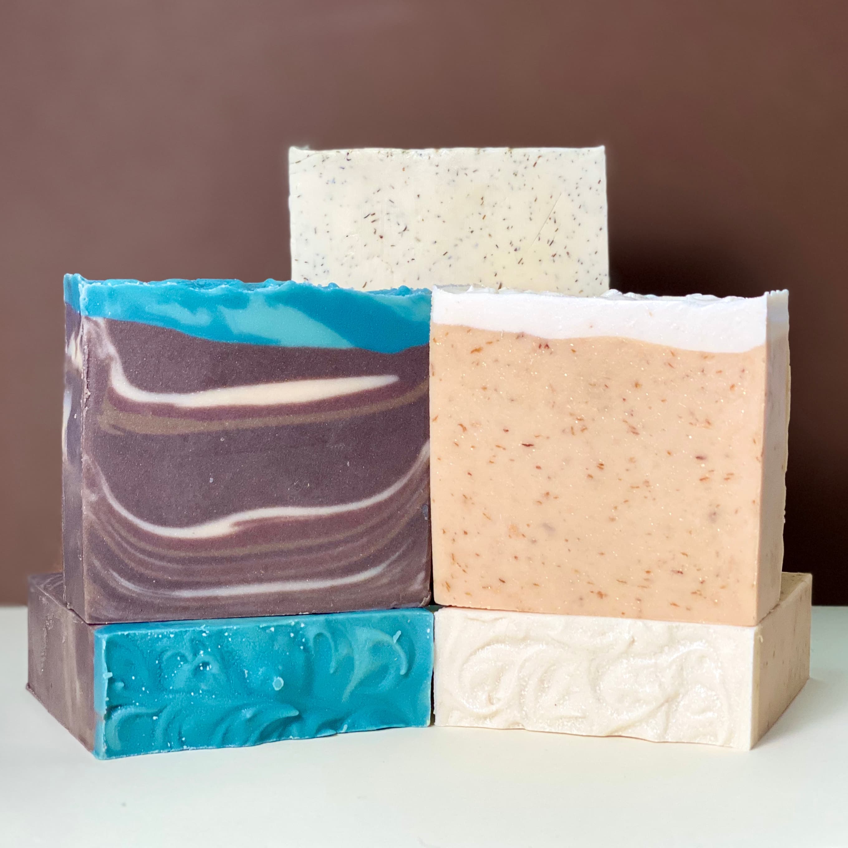 Six handcrafted soap bars stacked in a pyramid display. Colors include purple with blue swirls, peach with visible botanical flecks, cream with herbs, and white with specks throughout.