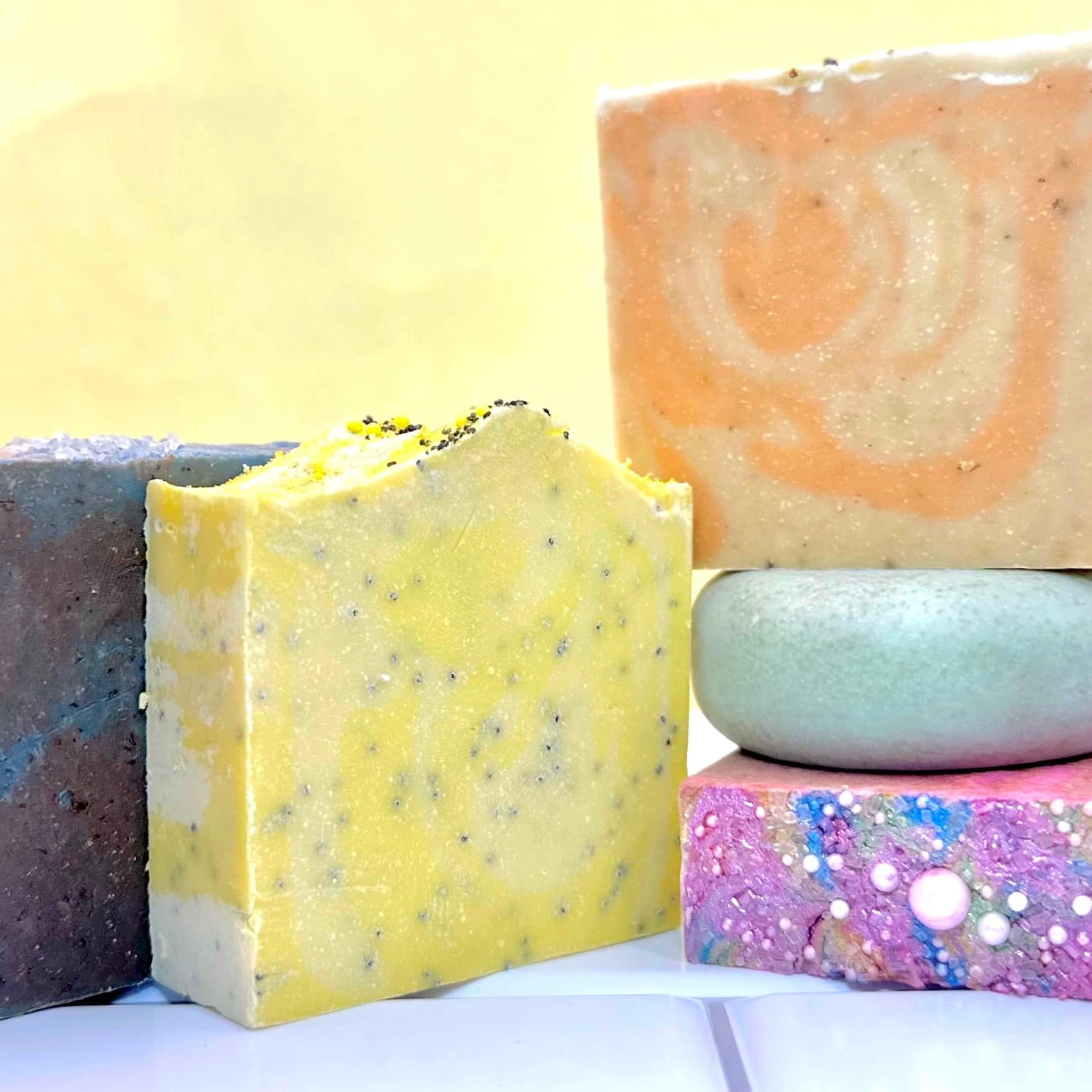 Five handcrafted soap bars arranged together displaying varied colors: yellow with poppy seeds, peach, purple-blue gradient, lavender with decorative bubbles, and dark brown-gray. Each bar shows distinct texture and exfoliating ingredients.