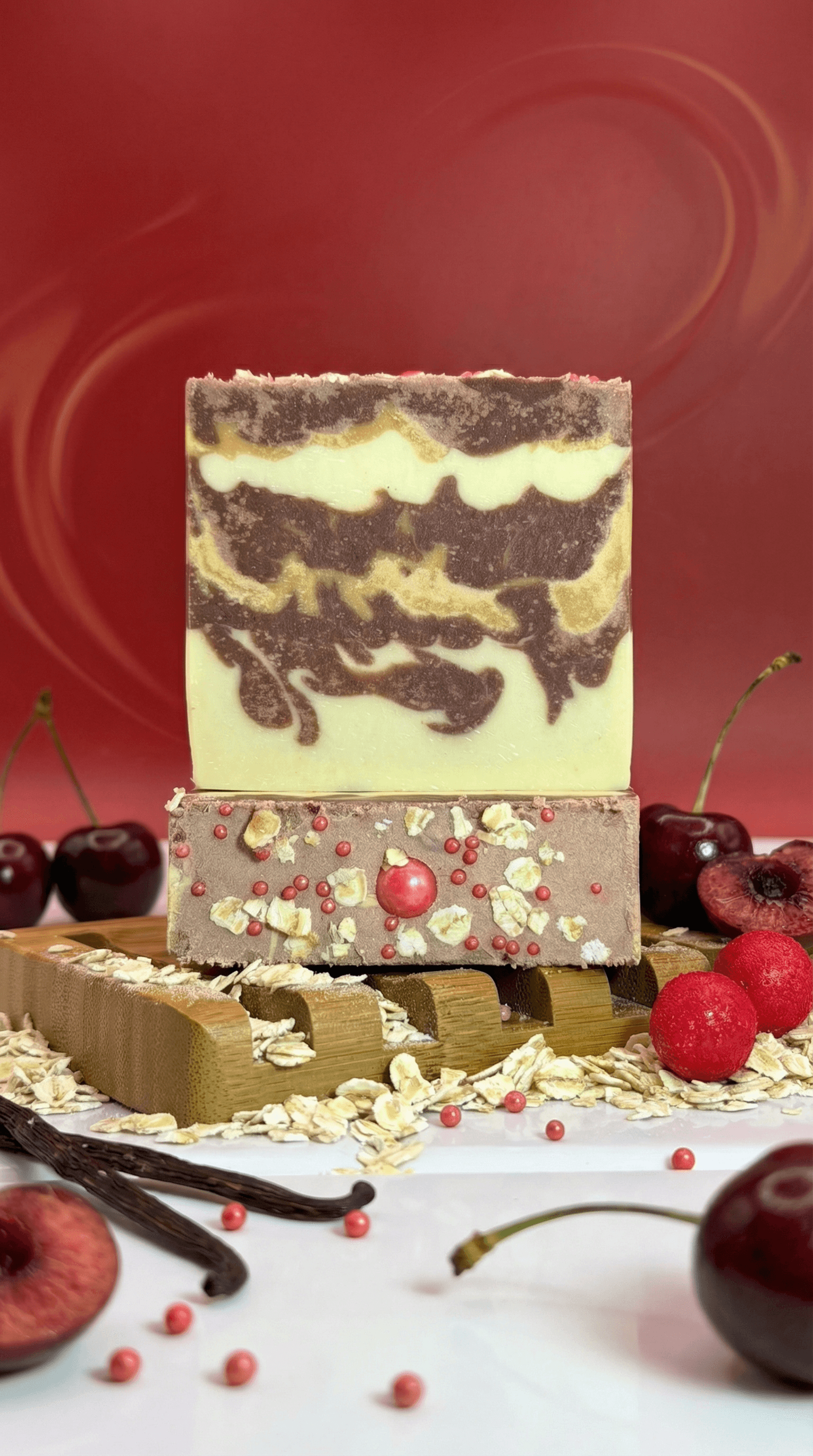 Two stacked handcrafted soap bars on a wooden holder surrounded by oats, cherries, peaches, vanilla beans, and pink peppercorns. Top bar features brown and cream swirled layers; bottom bar shows embedded fruit pieces and pink spheres in brown soap.