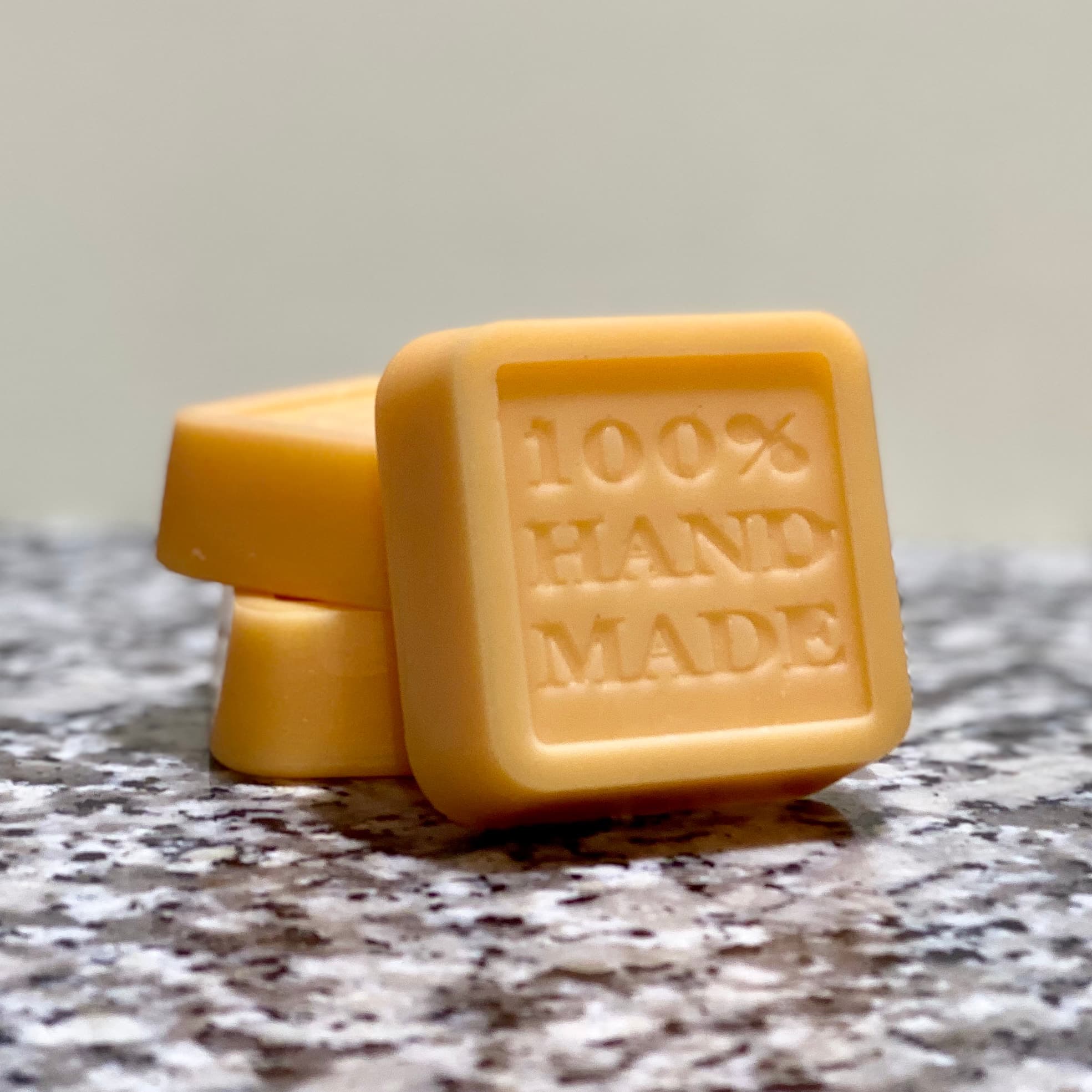 Three stacked golden yellow square bars with embossed text reading '100% HAND MADE' on speckled granite surface. Smooth, rounded edges with glossy finish.