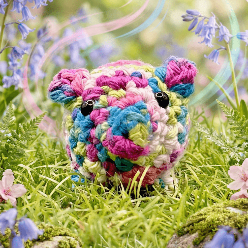 Colorful hand-crocheted pig sachet in pink, blue, yellow and white yarn sits in bright green grass surrounded by purple bluebells and moss, displaying detailed texture and black safety eyes.