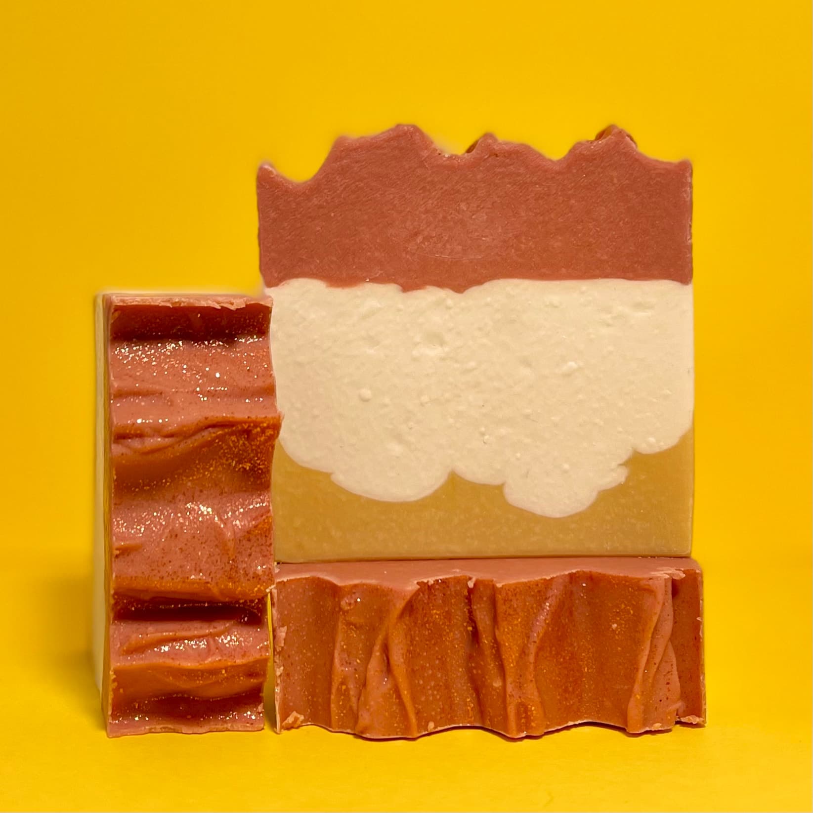 Three bars of handcrafted soap with distinct layers: rust-orange top, creamy white middle with scalloped edges, and golden-yellow bottom, displayed against a bright yellow background.