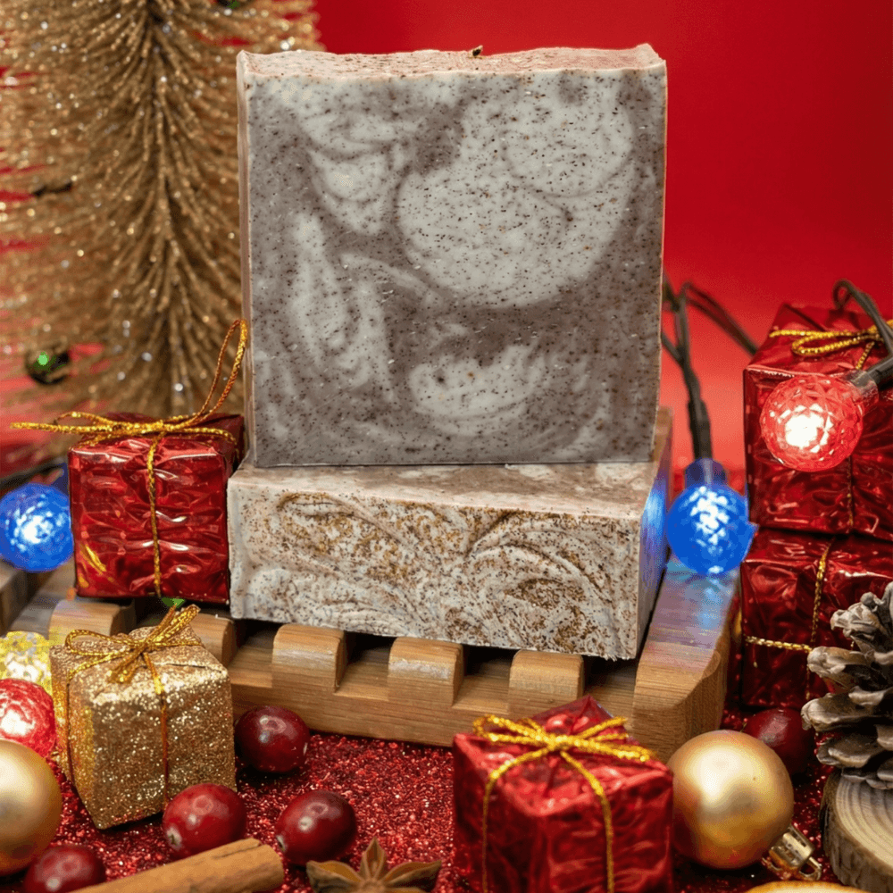 Two stacked handmade soap bars with brown and cream marbled swirls, displayed on a wooden soap dish surrounded by festive holiday ornaments, miniature gifts, and pine decorations.