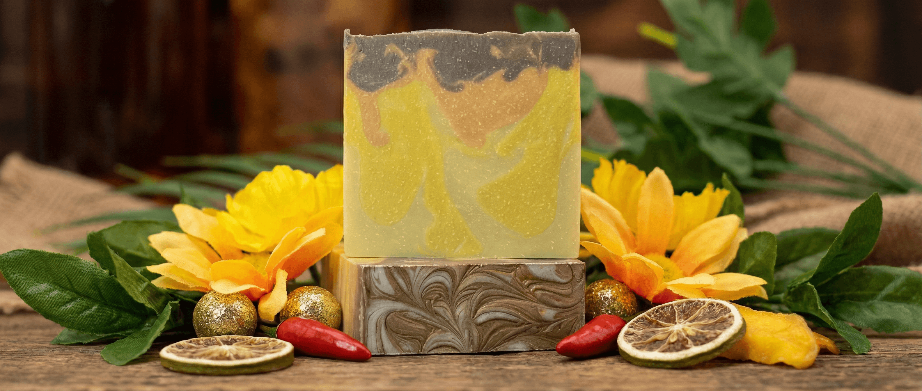 Handcrafted soap bar with swirled yellow, orange, and charcoal layers, displayed on decorative mold with dried citrus slices, yellow flowers, and golden ornaments on wooden surface.