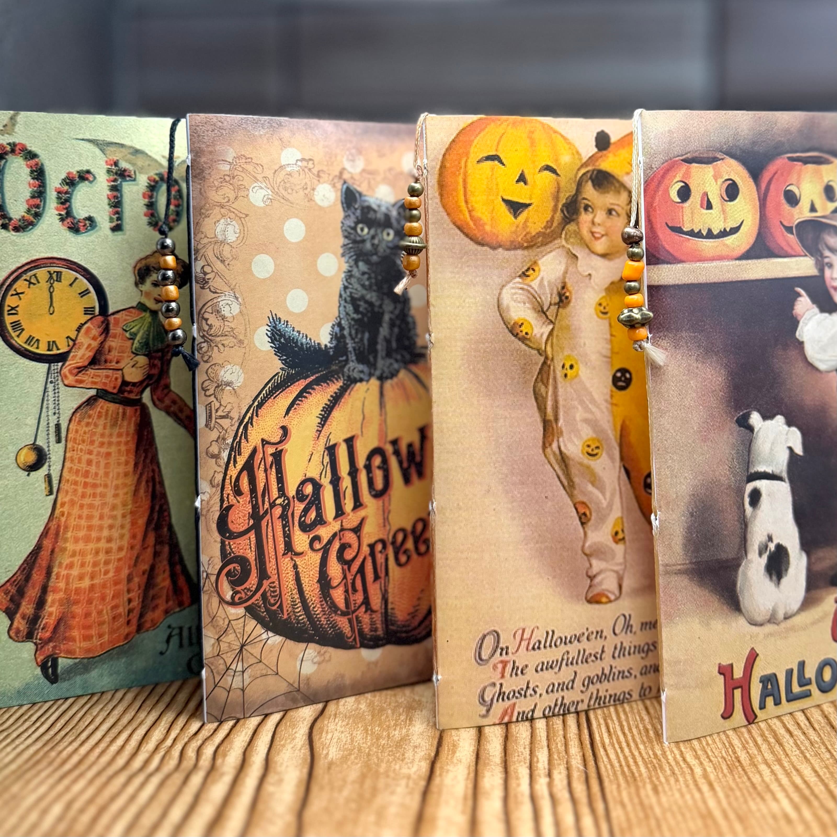 Four vintage-style Halloween greeting card journals standing upright in a row, featuring retro imagery of black cats, jack-o'-lanterns, and children in costumes. Each has decorative beaded cord binding along the spine.