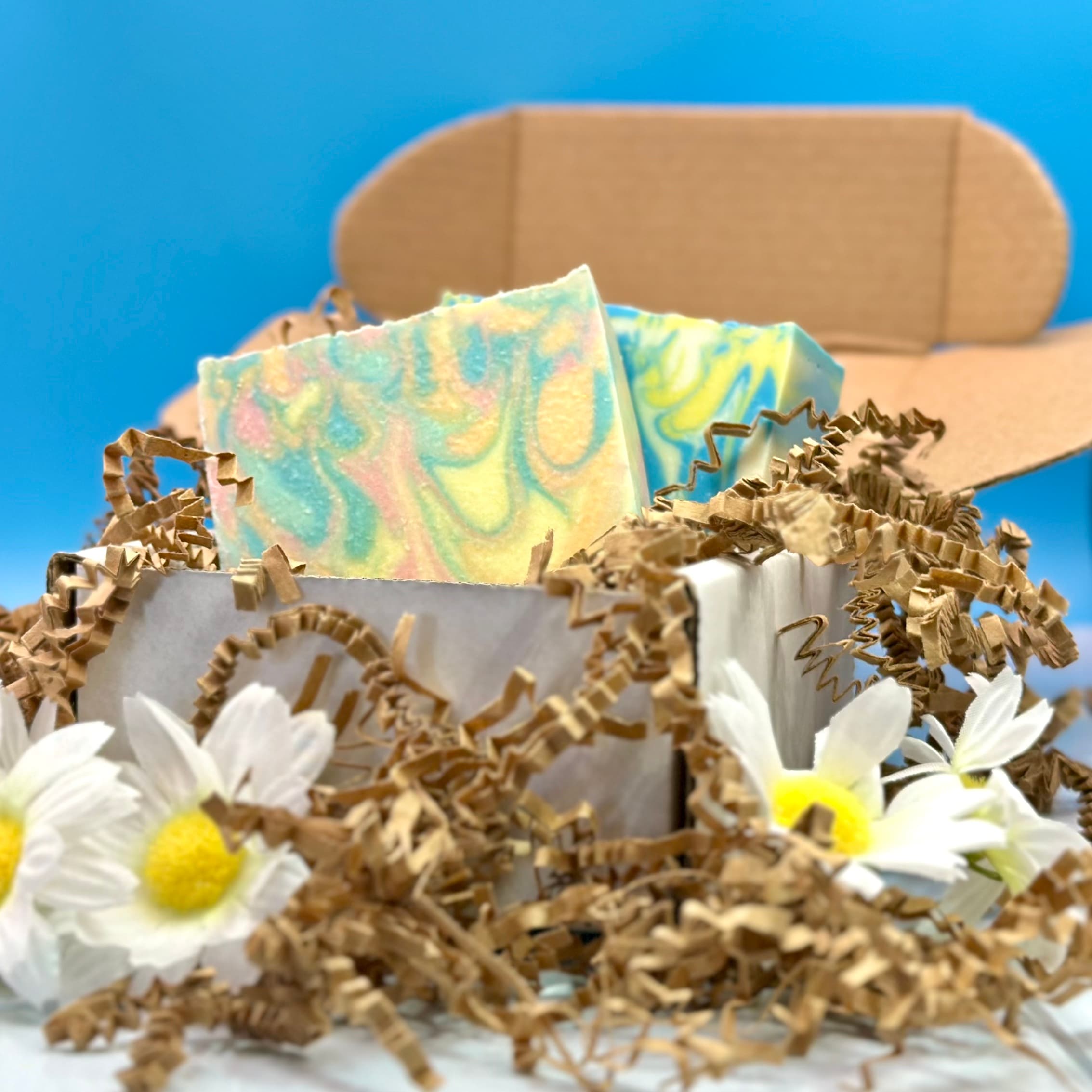 Colorful swirled soap bars in pastel yellow, blue, and pink nestle in kraft paper packaging with white daisies and crinkled natural paper shred against a bright blue background.