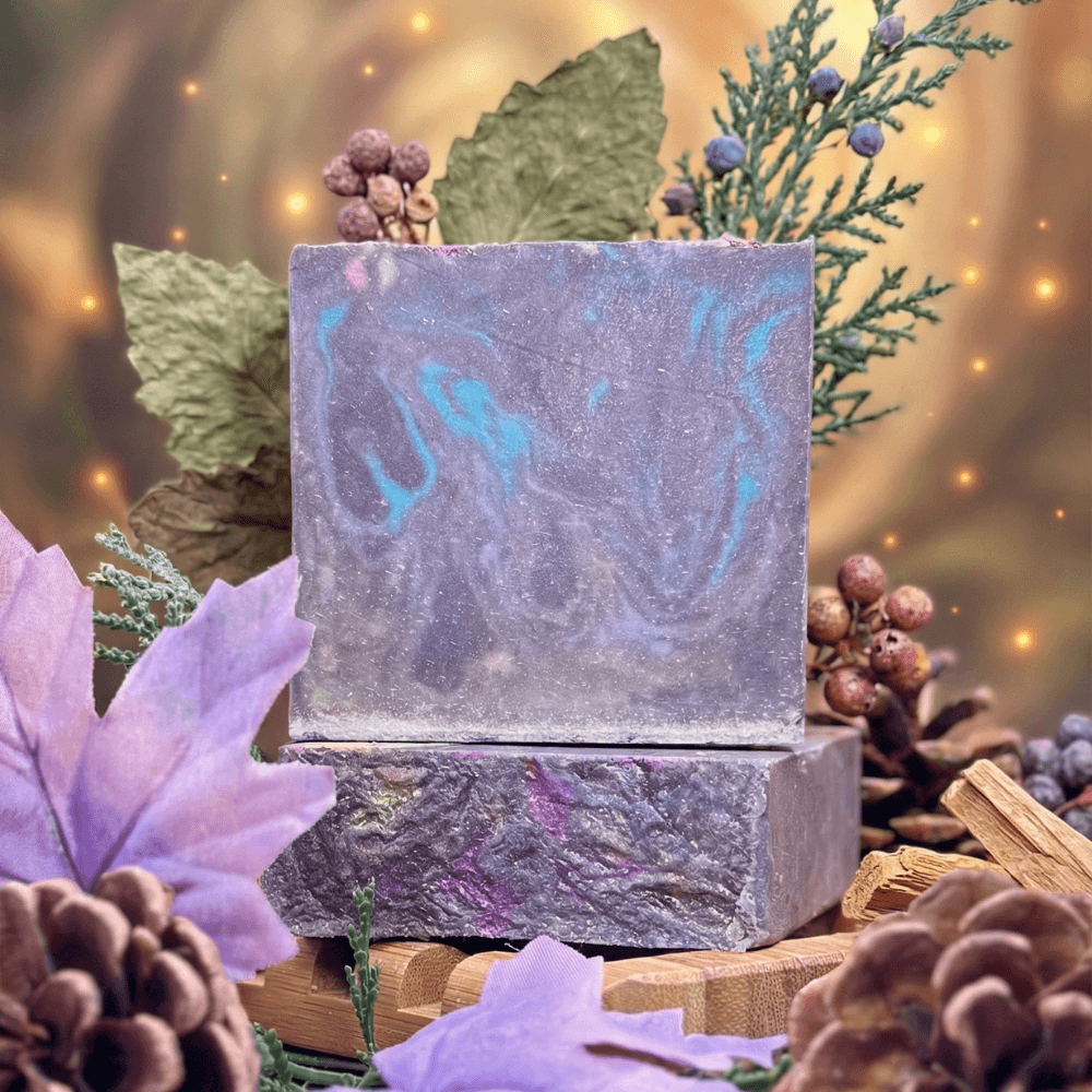 Two purple handmade soap bars with swirled turquoise accents, stacked on wood surrounded by dried botanicals, lavender flowers, berries, pine cones, and twinkling lights.