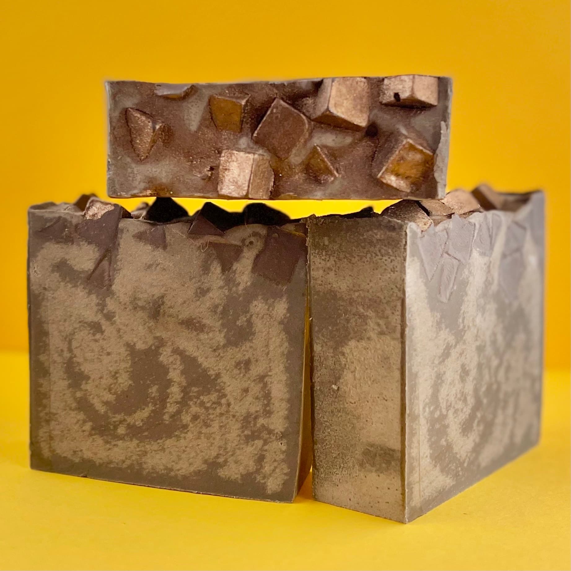 Two bars of brown and tan mottled soap stacked on bright yellow background. Top surfaces feature chunky amber and brown soap pieces embedded in surface.