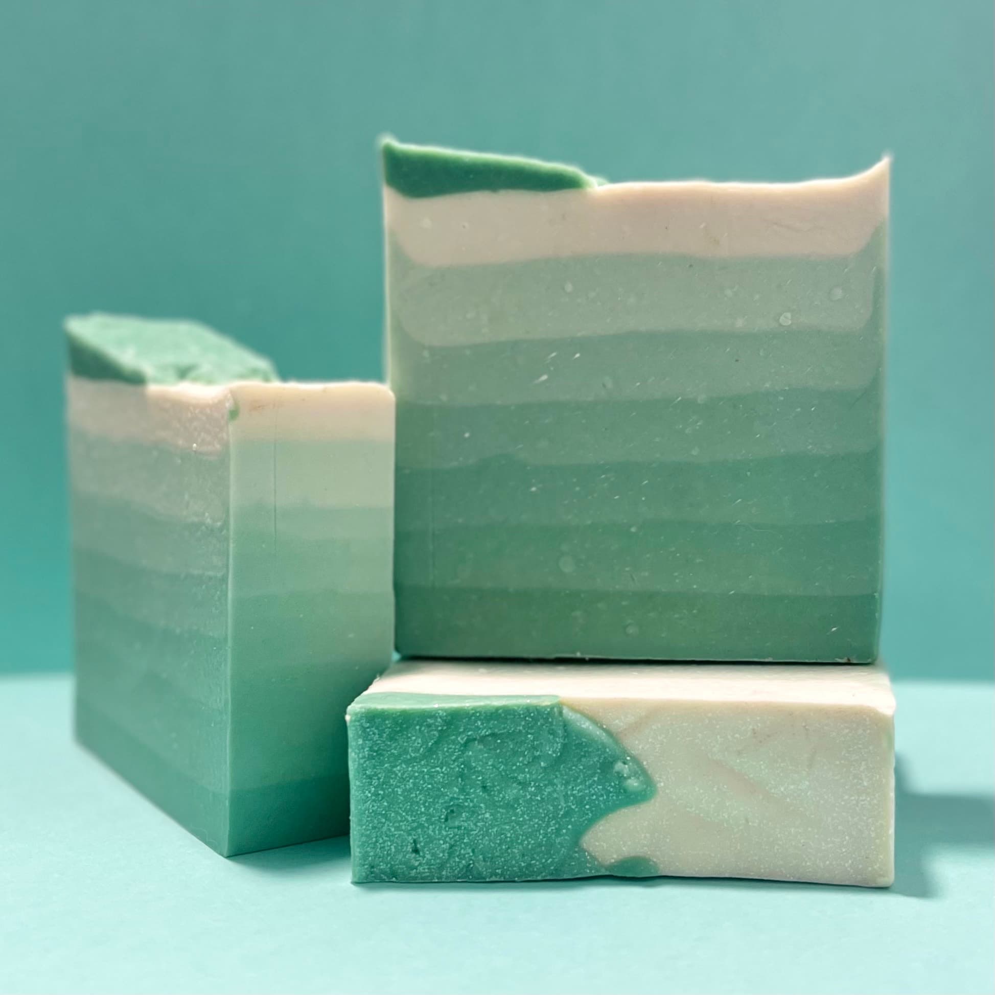 Three bars of handcrafted soap with diagonal teal and white swirls, featuring a smooth matte finish with subtle shimmer. Product-only display against a matching teal background.