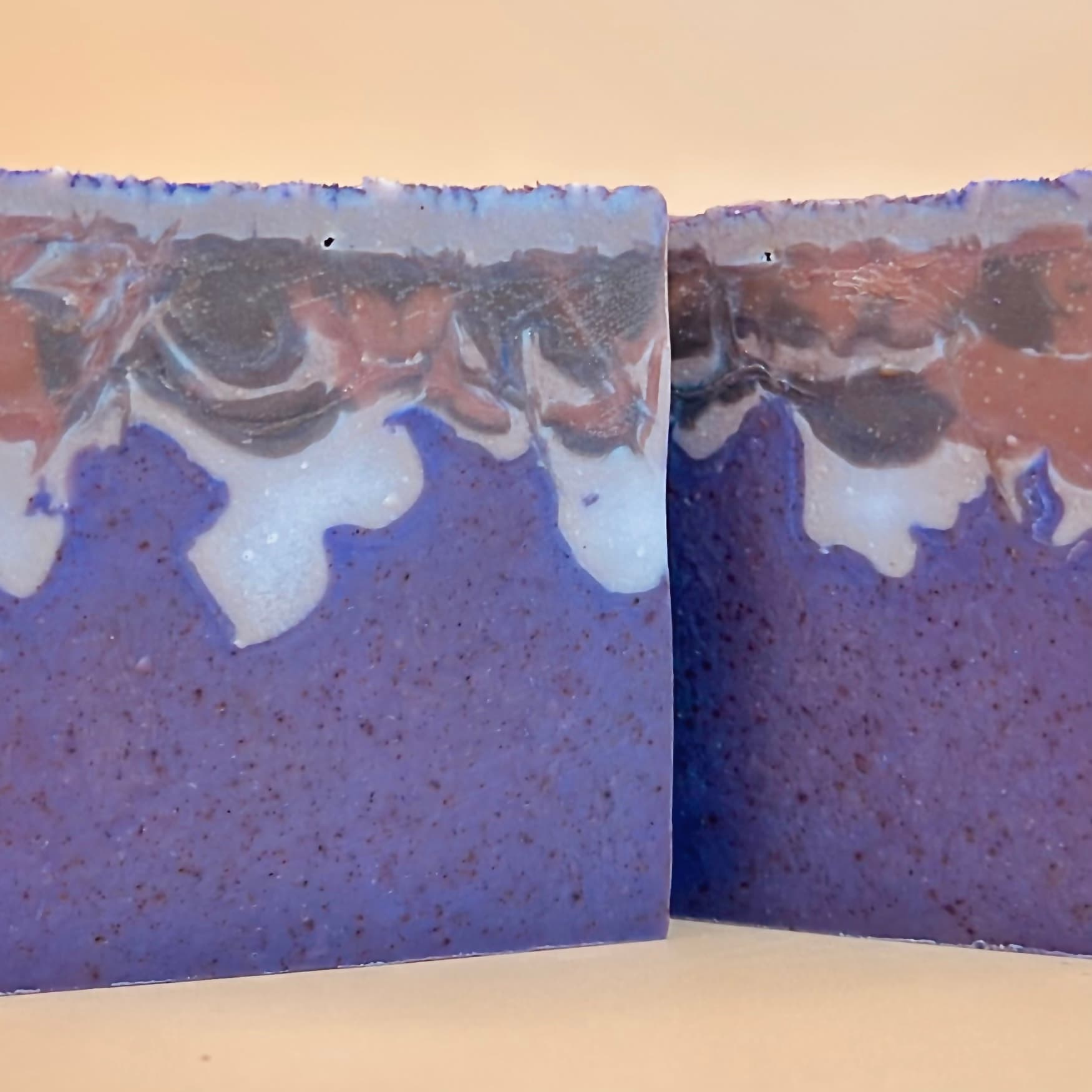 Two purple ombre soap bars with marbled white and rust-brown swirls on top. Darker speckles throughout indicate walnut shell exfoliant. Product-only view against neutral background.