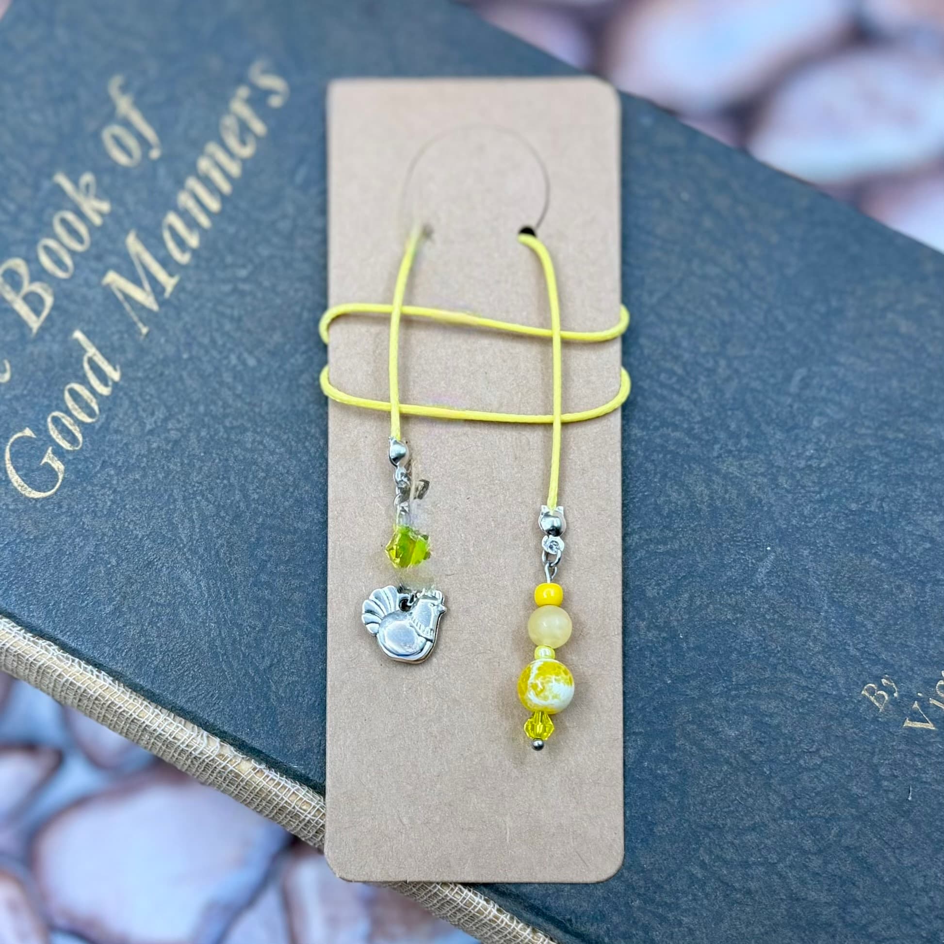 Two beaded bookmarks with yellow cord attached to a kraft card, resting on a dark book titled 'Book of Good Manners'. One features a silver hen charm, the other has yellow and cream beads.