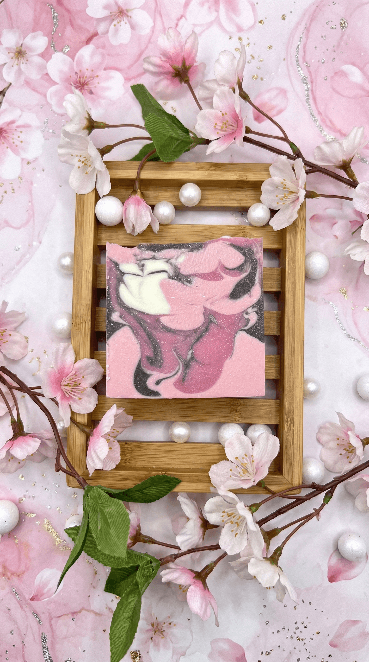 Handcrafted soap bar with pink, white, and charcoal gray marbled swirls on bamboo tray, surrounded by cherry blossoms and pearl accents on soft pink floral background.
