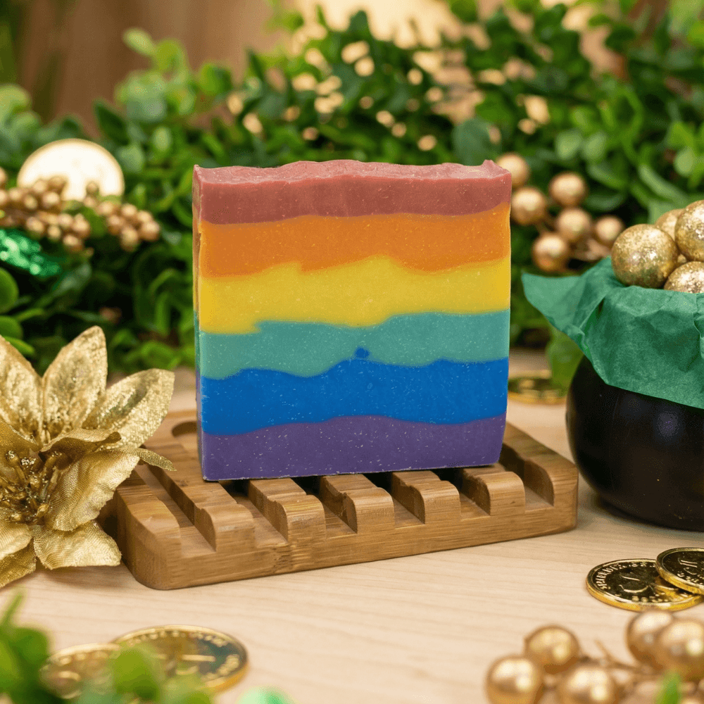 Rainbow-layered handmade soap bar with six vibrant horizontal stripes from purple to pink, resting on a wooden soap dish surrounded by gold decorative elements and greenery.