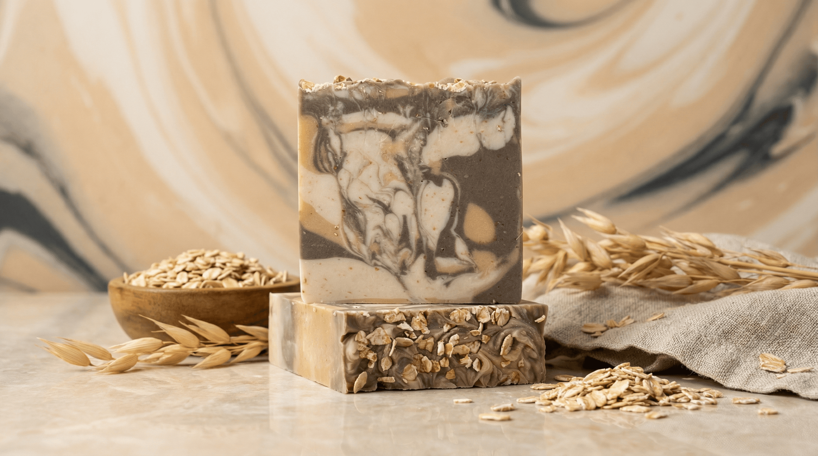 Two handcrafted soap bars with brown and cream marbled swirls, topped with whole oats. Displayed with scattered oat flakes, wheat stalks, and wooden bowl on neutral linen backdrop.