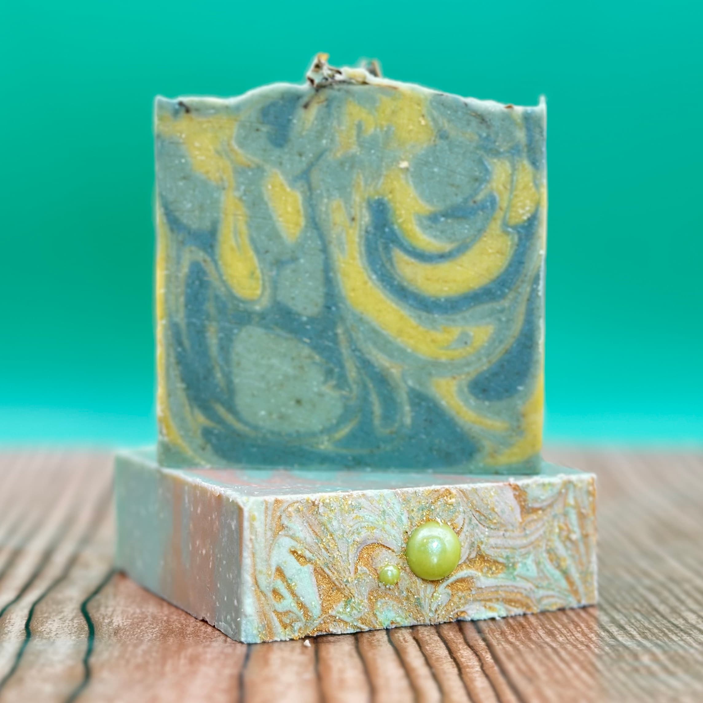Two stacked handcrafted soap bars on wood surface against turquoise background. Top bar features blue-gray and yellow swirled pattern, bottom bar has gold shimmer swirls with two lime-green pearl embellishments.