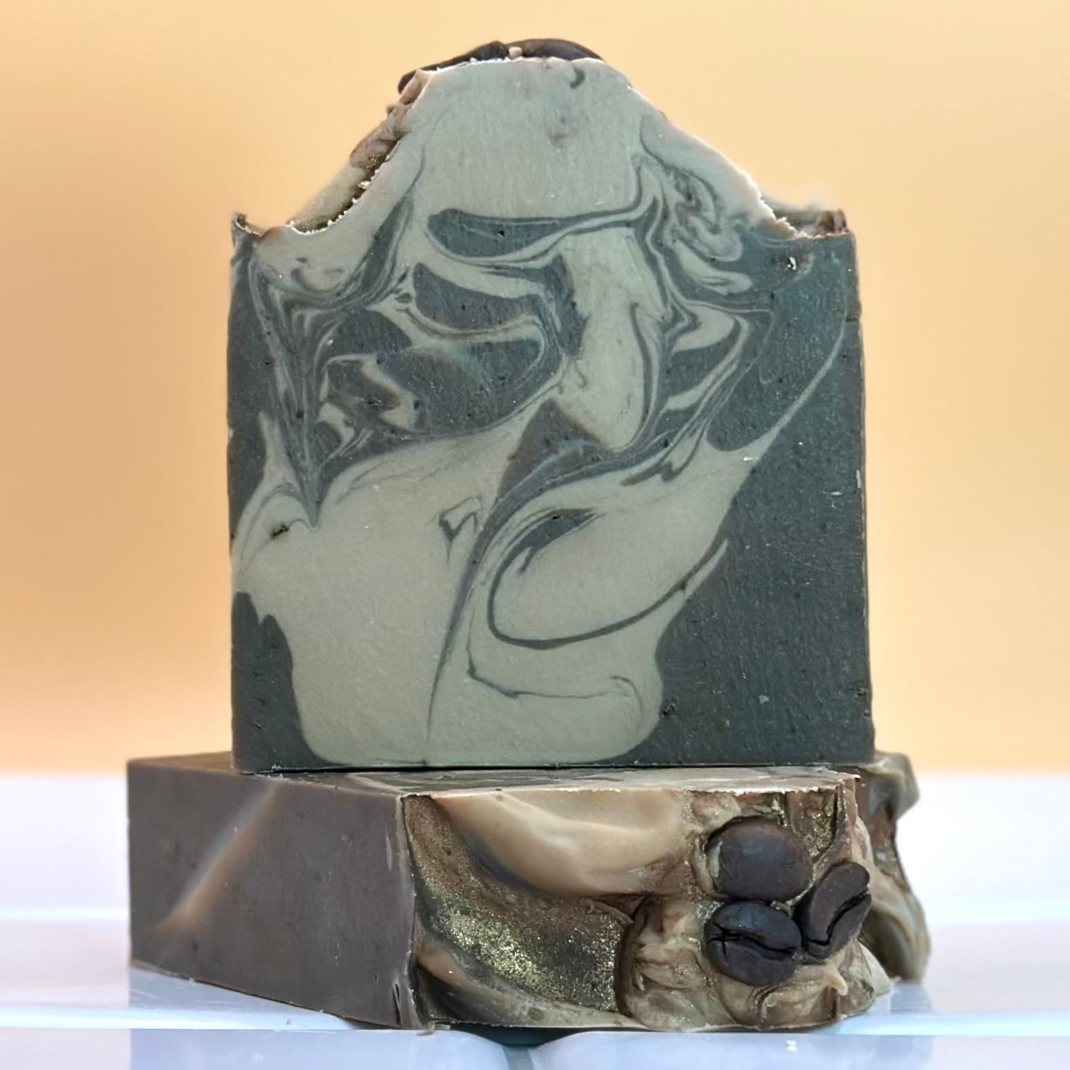 Two stacked handcrafted soap bars with marbled swirls of light and dark gray. The top bar features flowing marble patterns while the bottom has metallic gold accents and coffee bean decorations.