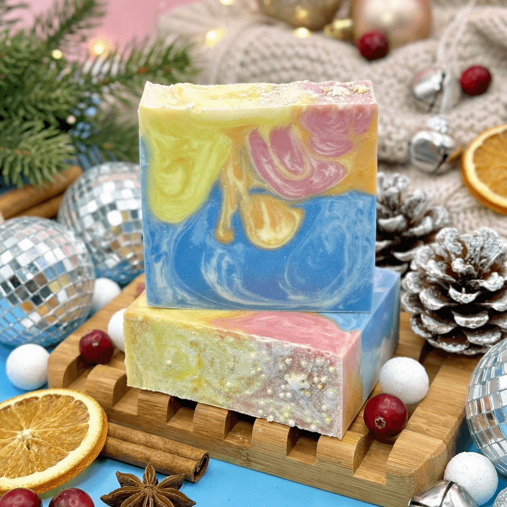 Two stacked artisan soap bars with swirled yellow, pink, and blue marbling on a wooden soap dish. Lifestyle styling includes disco balls, frosted pine cones, dried citrus, and holiday decorations.