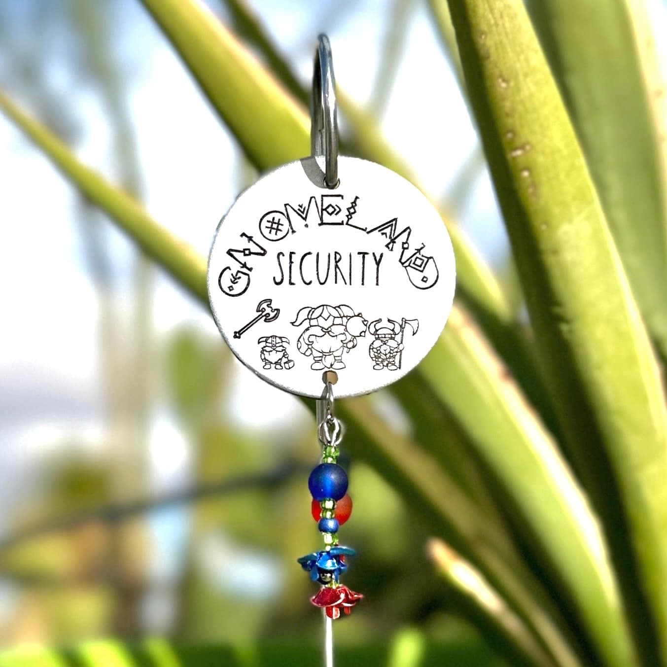 Round white aluminum disc hanging from a metal hook with hand-stamped text reading 'SECURITY' surrounded by gnome illustrations, decorated with blue and red glass beads dangling below, displayed among green plant stems.
