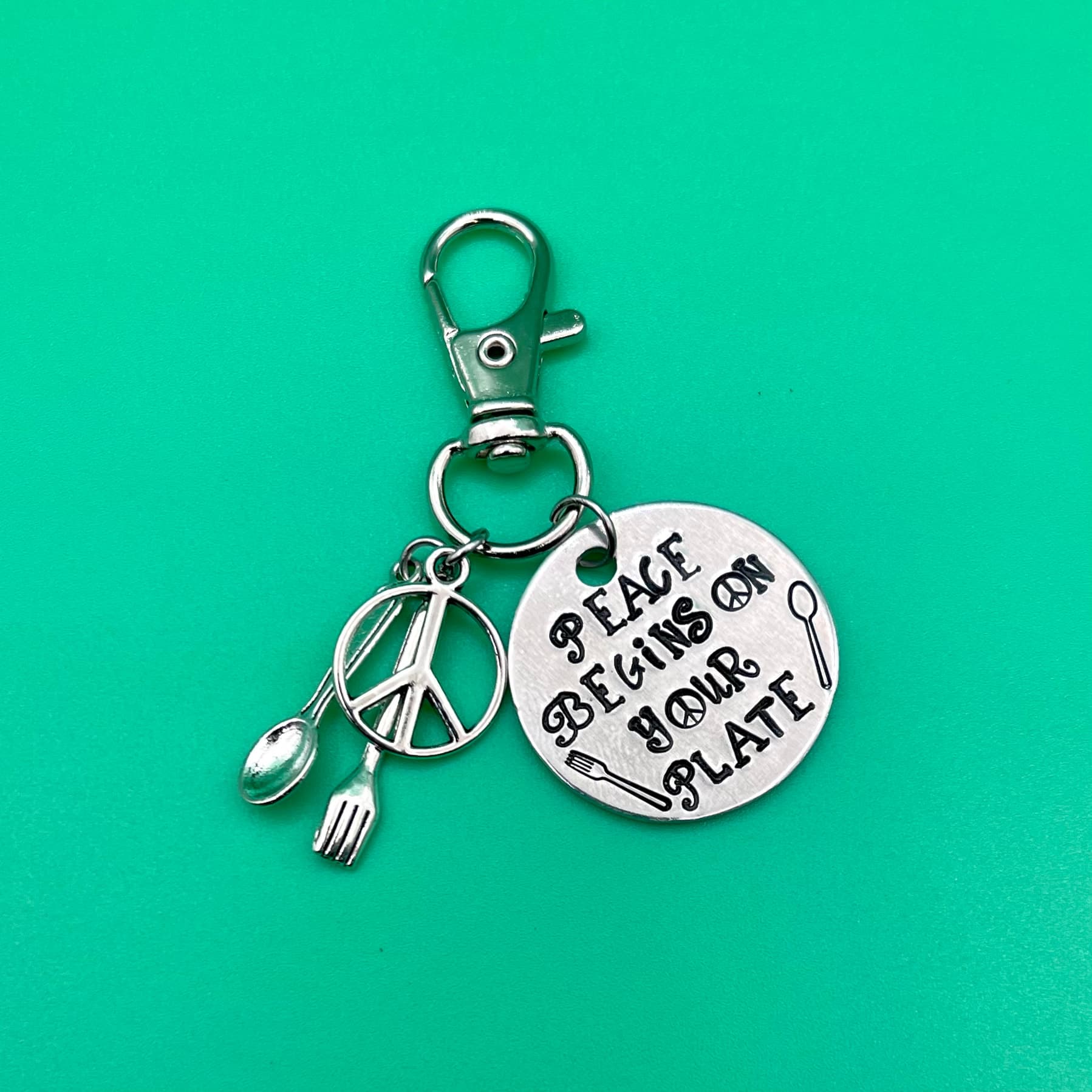 Silver keychain charm set on bright turquoise background featuring round stamped disc with peace symbol, fork, spoon, and utensils charm attached to lobster clasp.