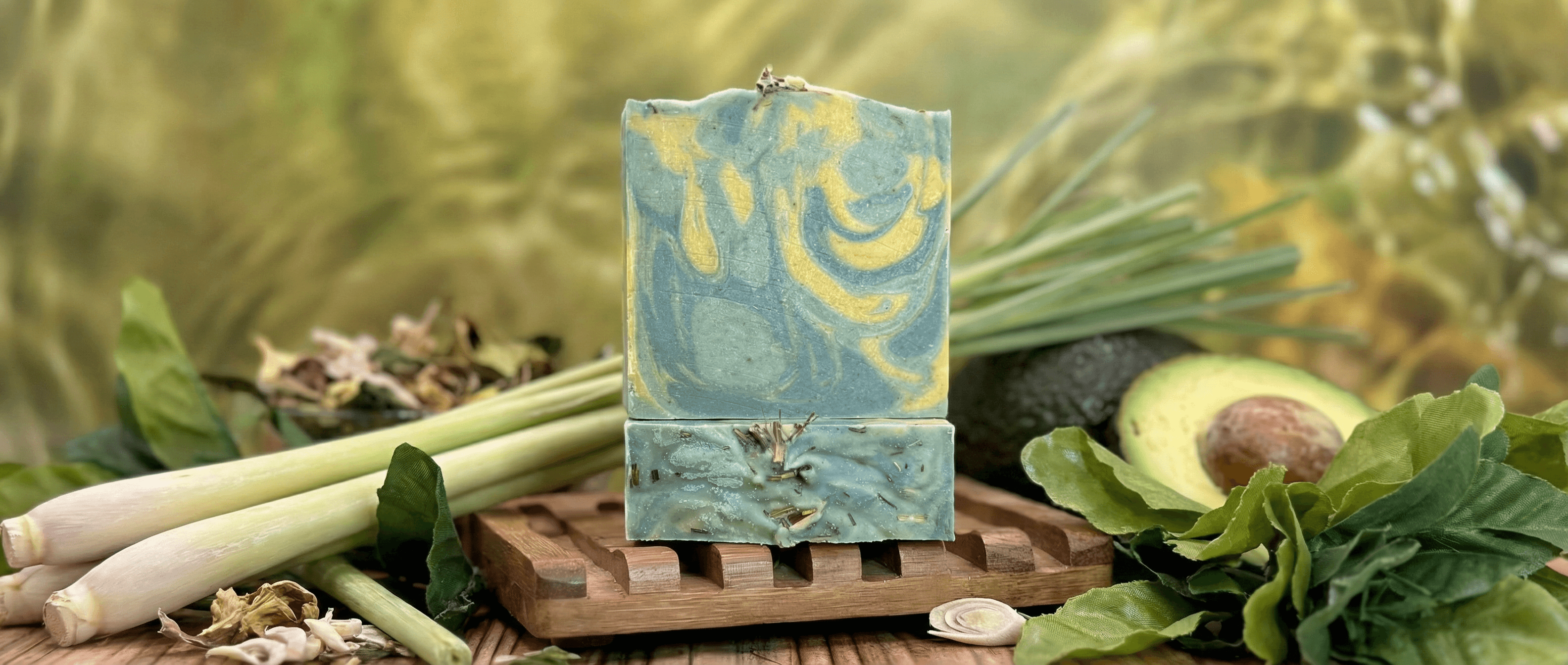 Blue and yellow marbled soap bar with dried botanicals embedded in the base, displayed on a wooden soap dish surrounded by fresh lemongrass stalks, avocado, and green leaves.