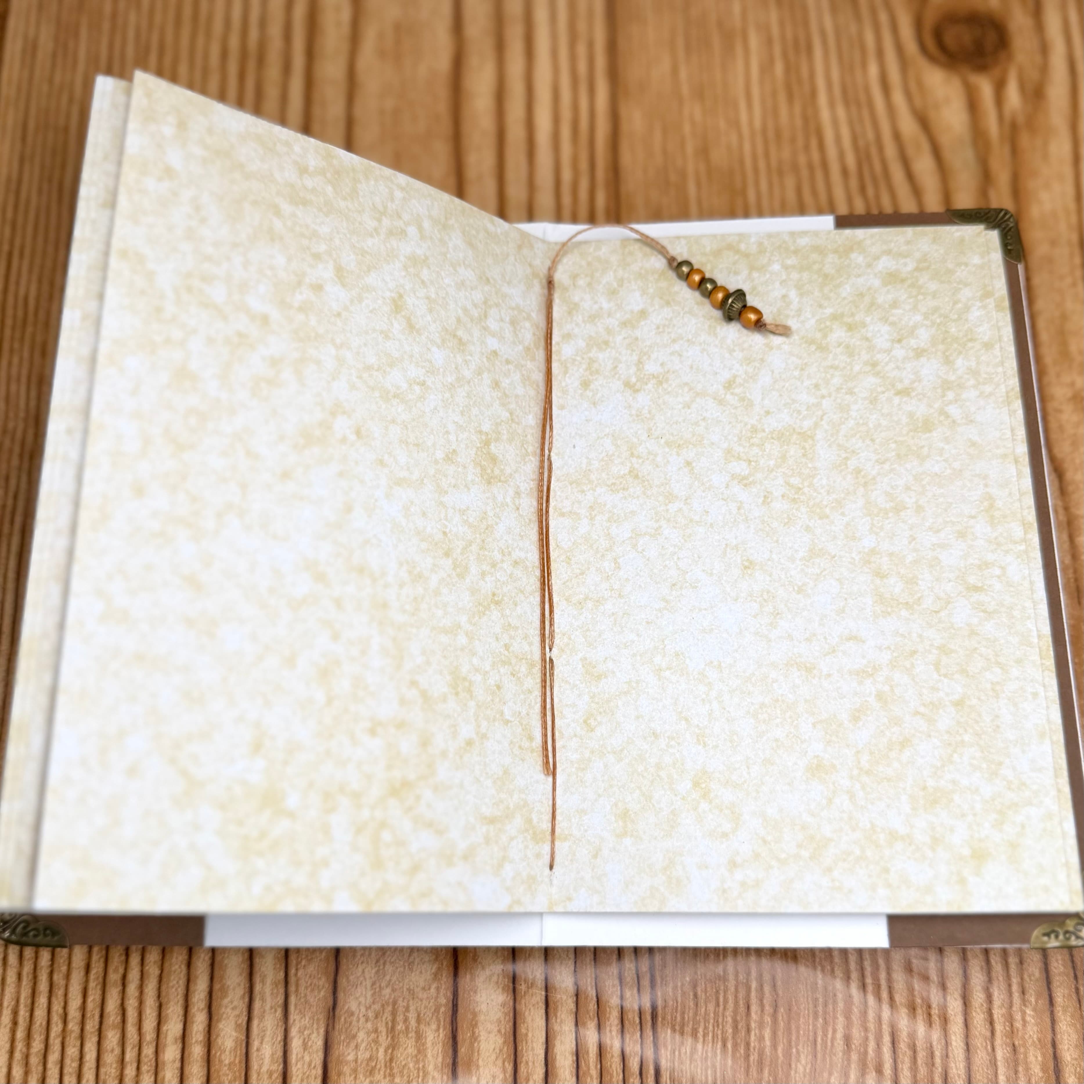 Open journal with blank cream-colored textured pages and beaded tan bookmark, displayed on wooden surface. Journal features metal corners and vintage aesthetic binding.