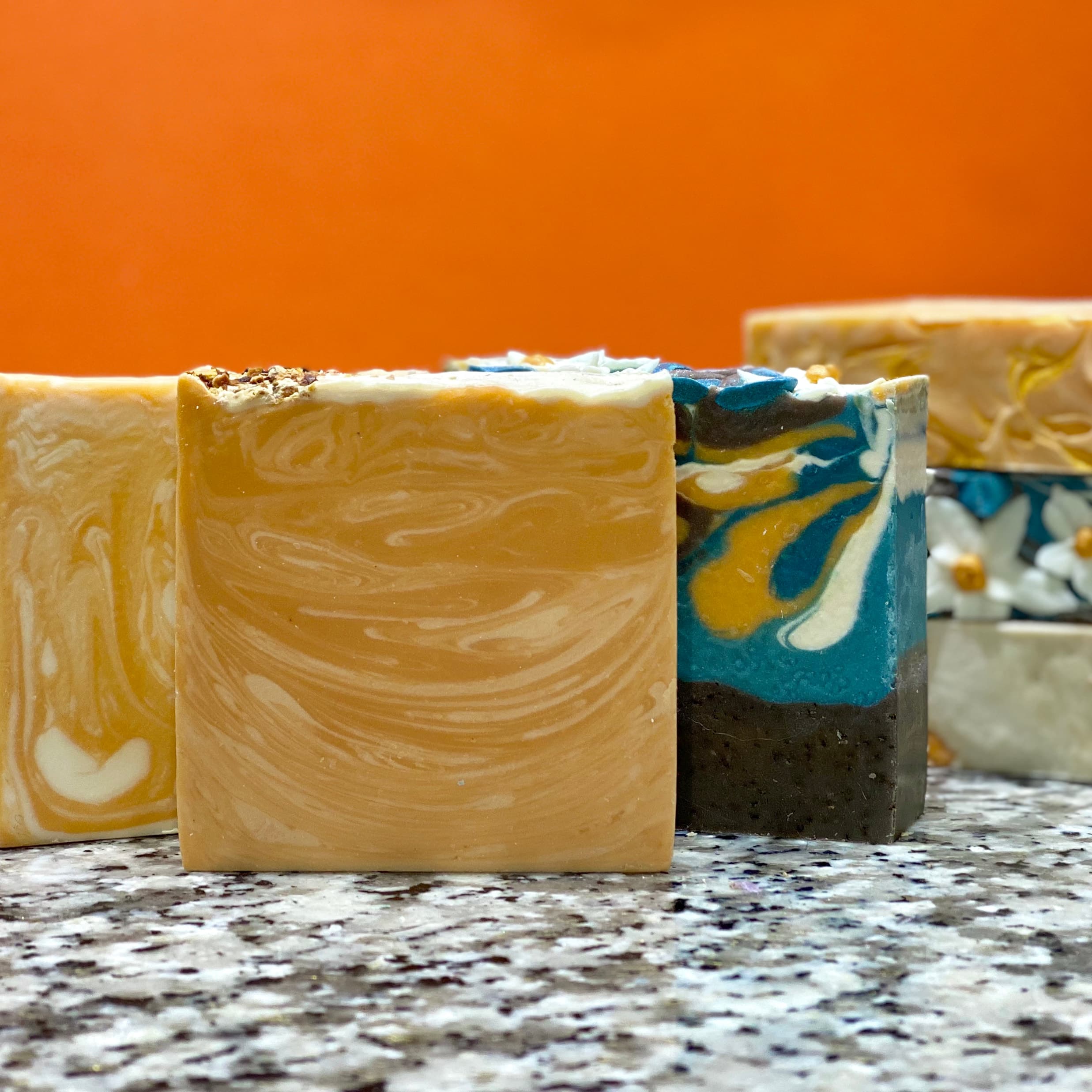 Four bars of handcrafted soap on speckled granite. Three feature swirled yellow-orange and white patterns; one has teal, yellow and charcoal marbled layers. Orange background.