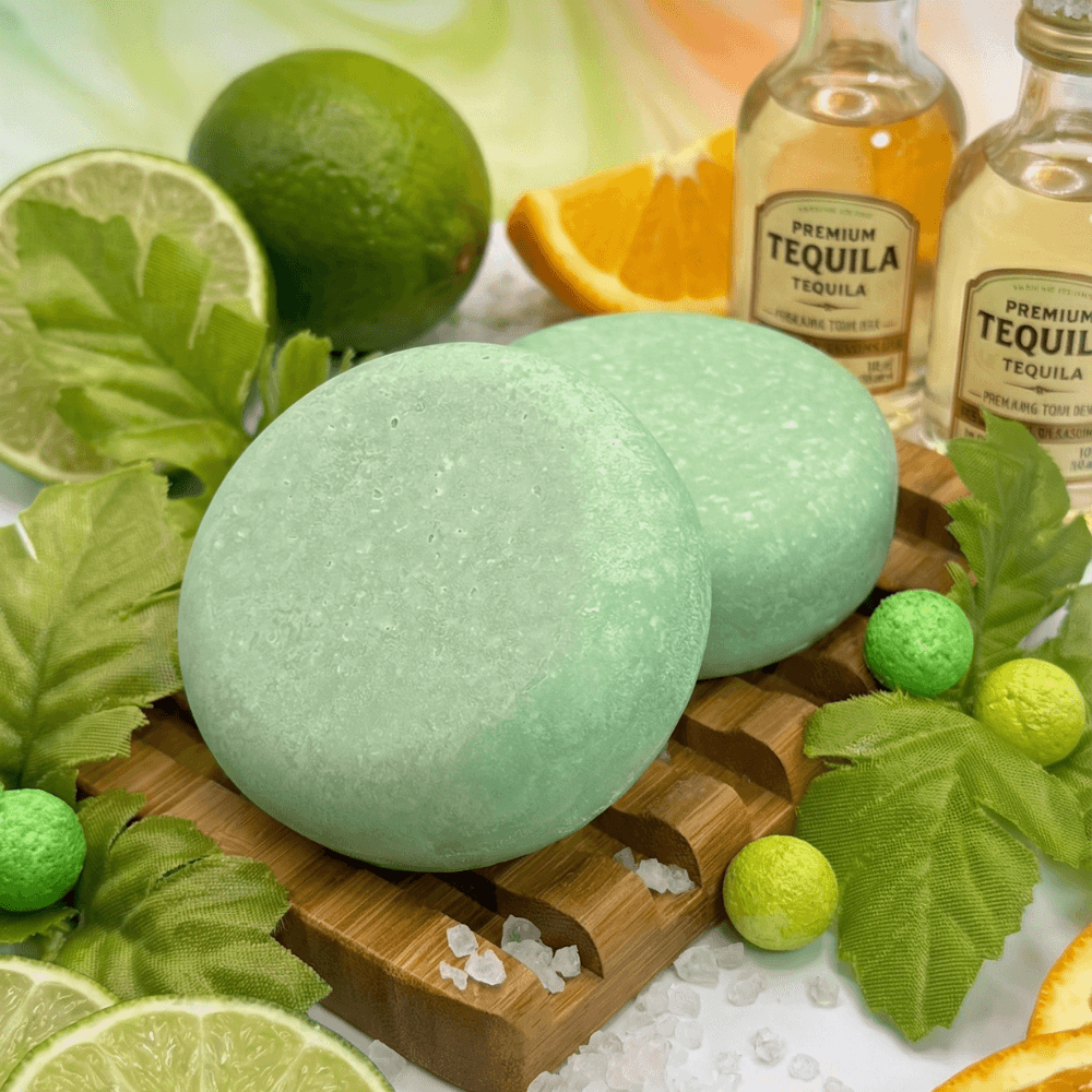Two pale seafoam green round soap bars with a subtly sparkled surface, displayed on a wooden board surrounded by fresh limes, orange slices, tequila bottles, and decorative salt crystals.