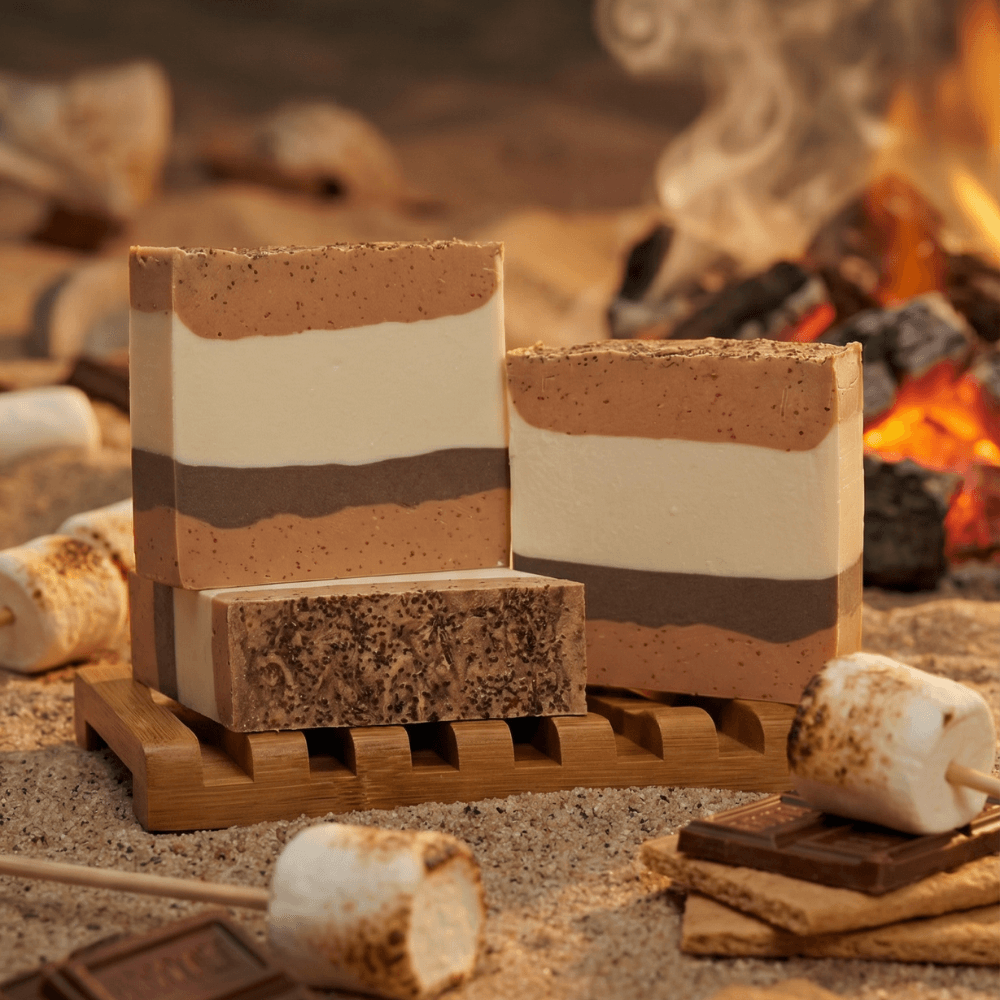 Three layered soap bars in brown, cream, and tan stripes with speckled texture, displayed on a wooden soap dish. Styled with toasted marshmallows, graham crackers, and chocolate by a glowing fire.