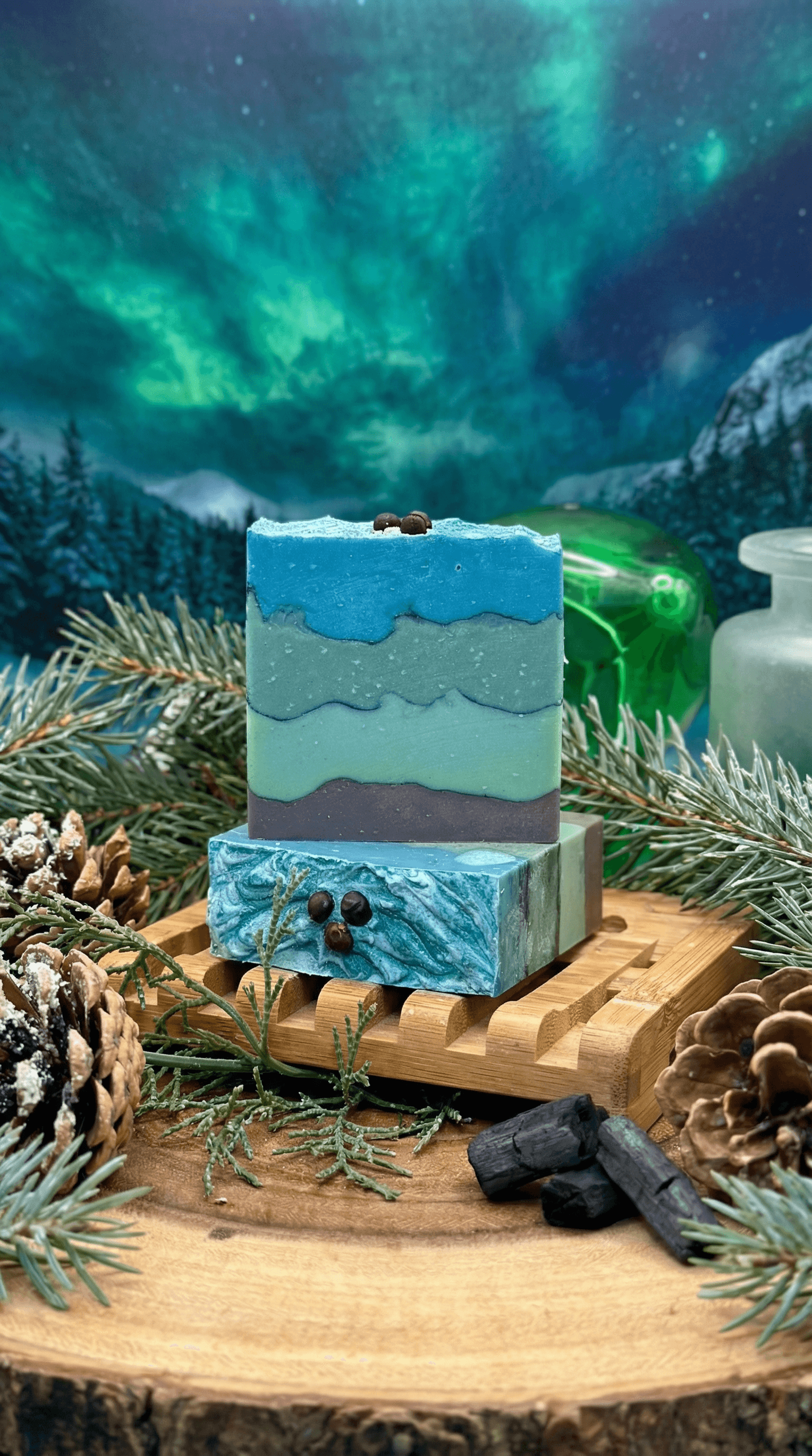 Layered handmade soap bar with wavy horizontal stripes in blue, teal, mint, and charcoal gray tones, displayed on wooden soap dish surrounded by pine branches, pinecones, and charcoal pieces with northern lights backdrop.