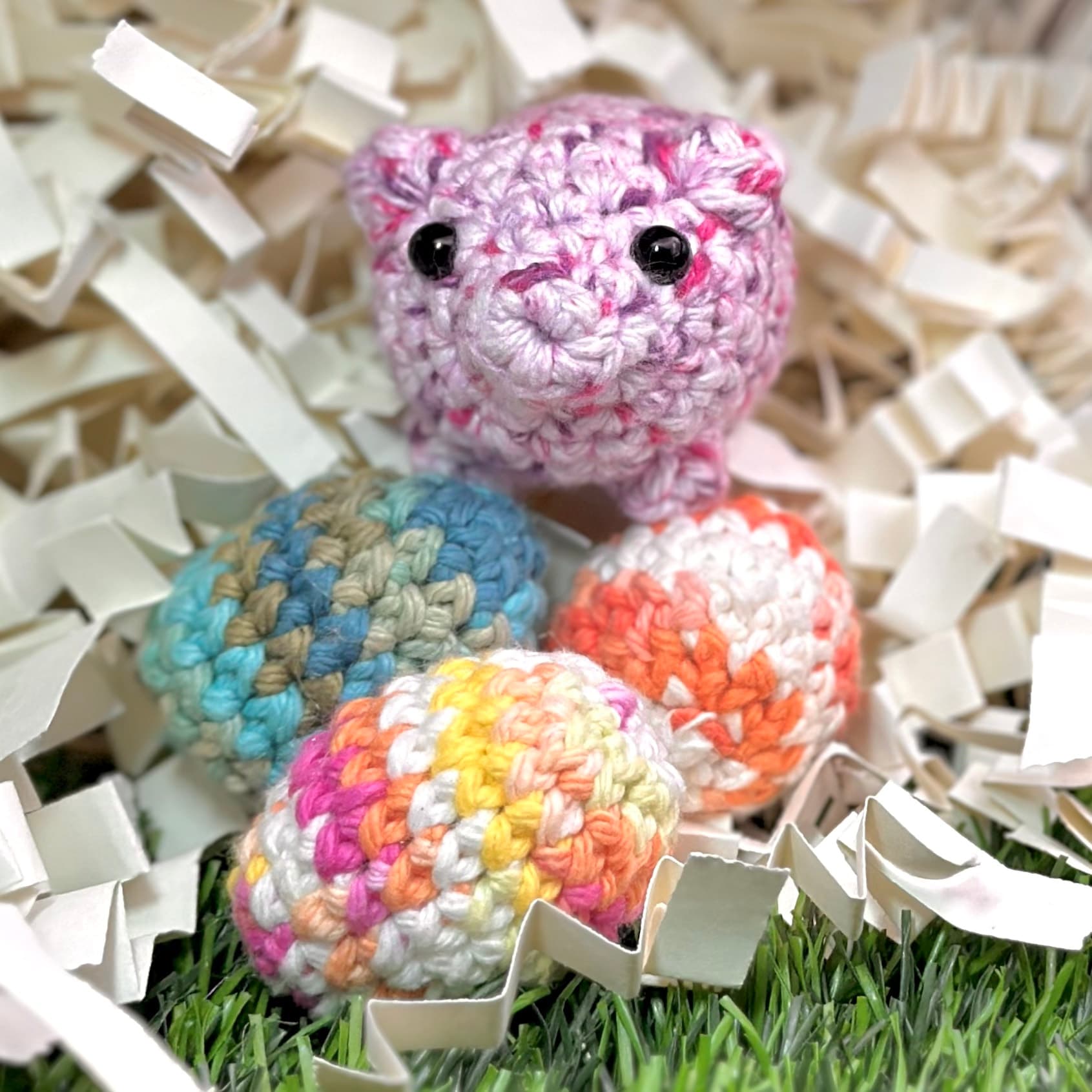 Four crocheted eggs in pastel colors arranged on artificial grass with white paper shred confetti. The pink speckled egg on top has a smiling face with black bead eyes.