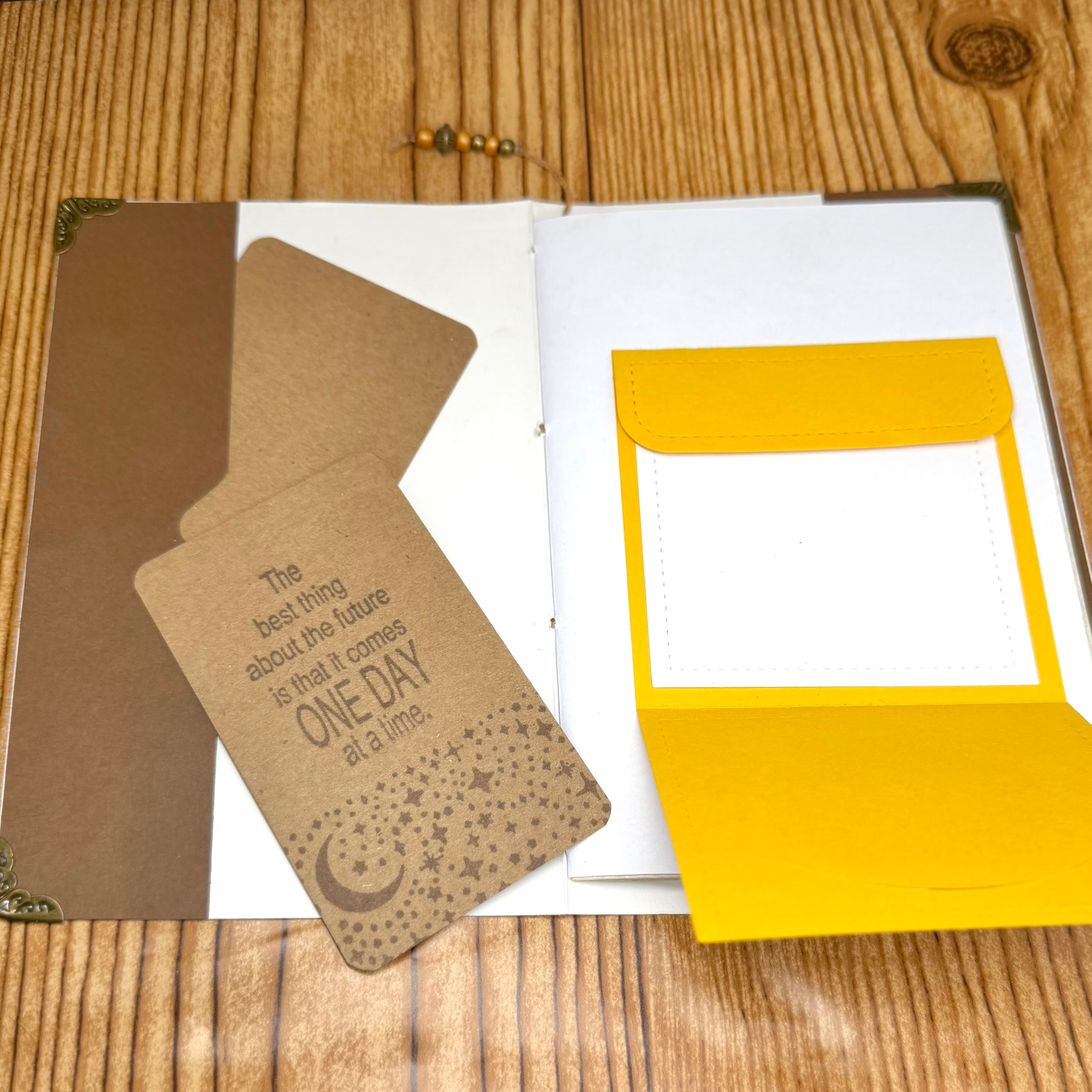 Flat lay of kraft brown journal with inspirational quote and yellow pocket-style journal, both with blank affirmation cards, displayed on wooden surface with metal corner details visible.