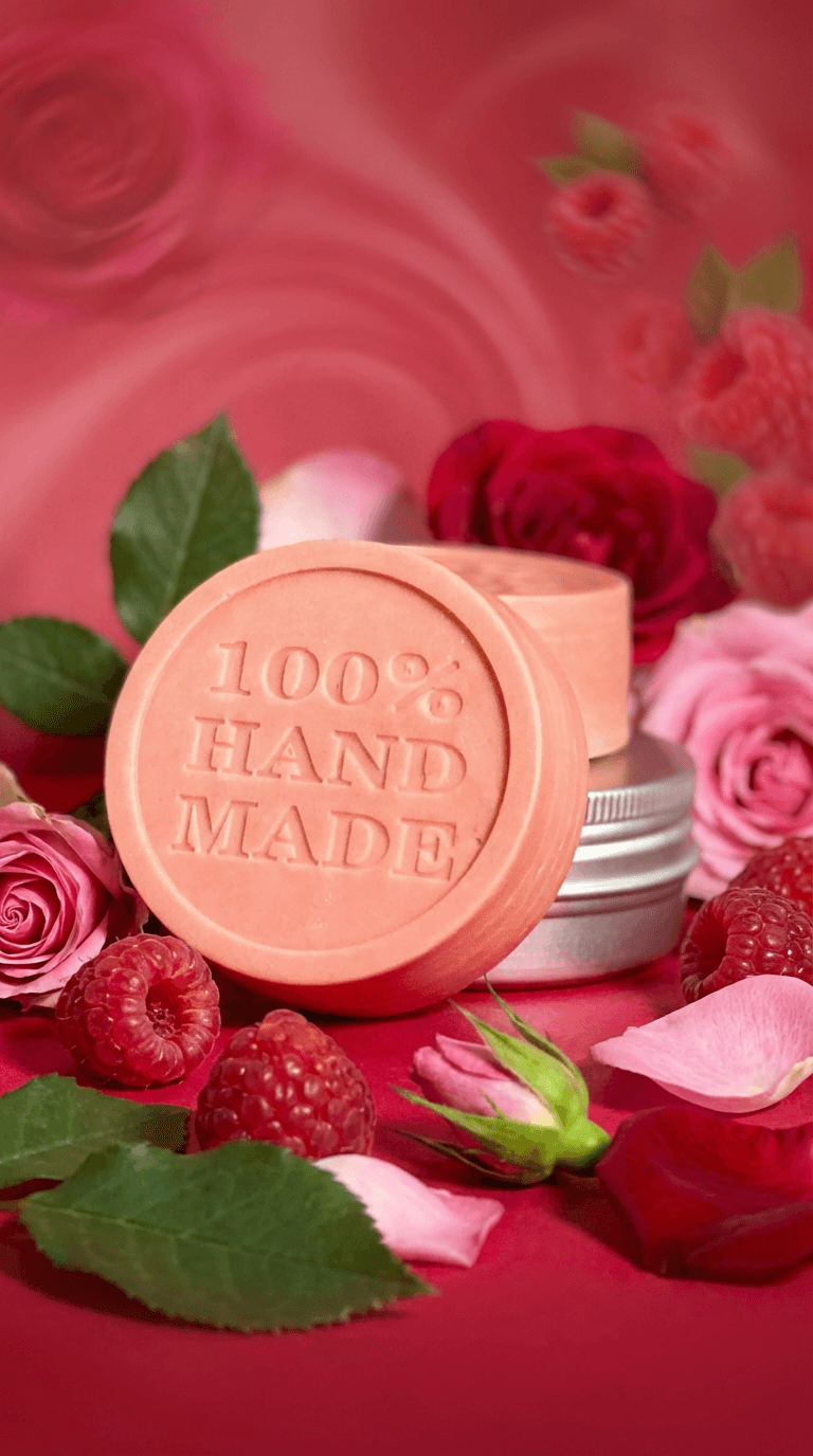 Peachy-pink solid lotion bar stamped with '100% HAND MADE' displayed in aluminum tin, surrounded by fresh roses, rose petals, raspberries and green leaves on pink background.